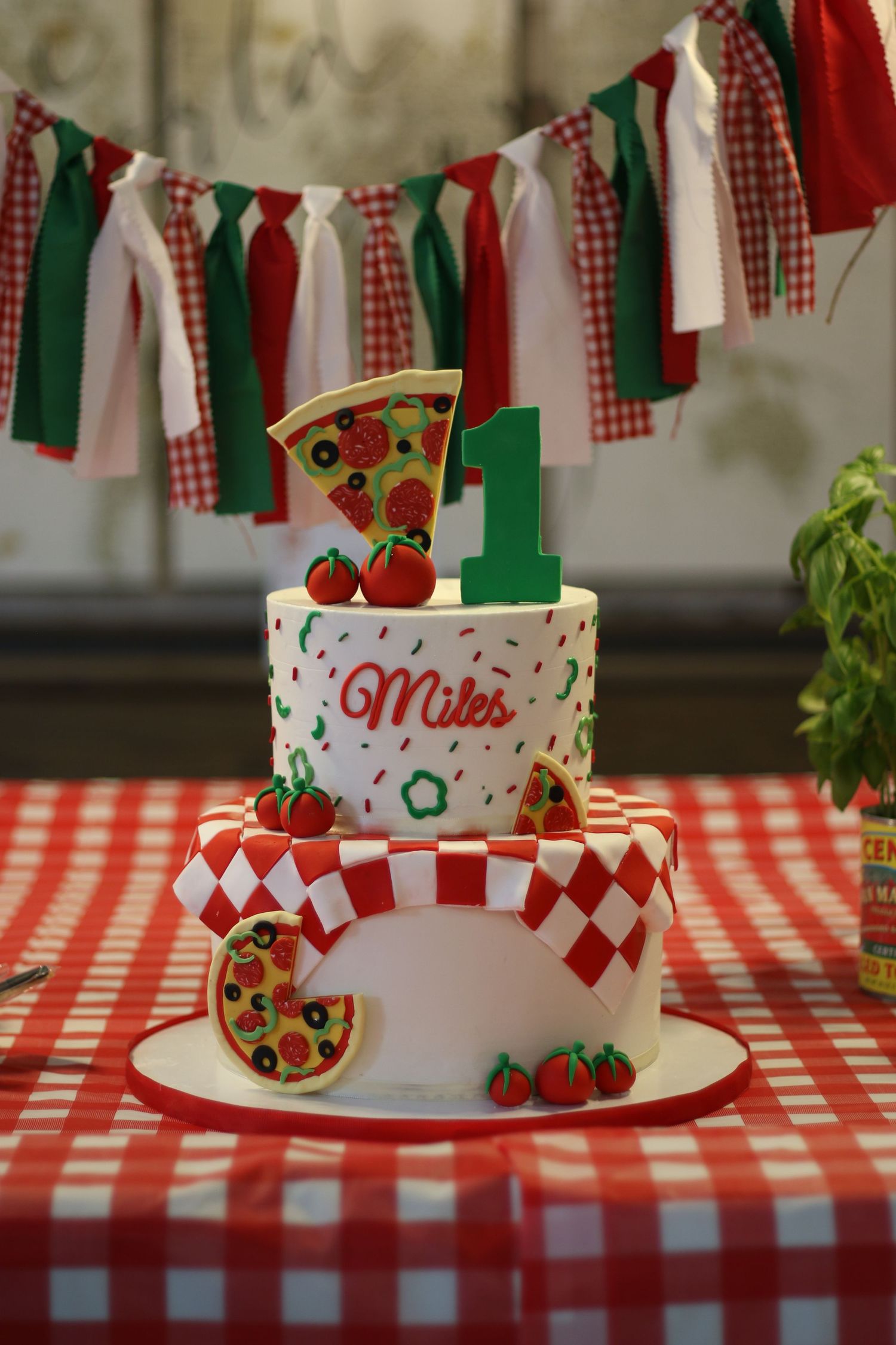 Two-tier pizza-themed first birthday cake decorated with the name Miles on a red checkered tablecloth.