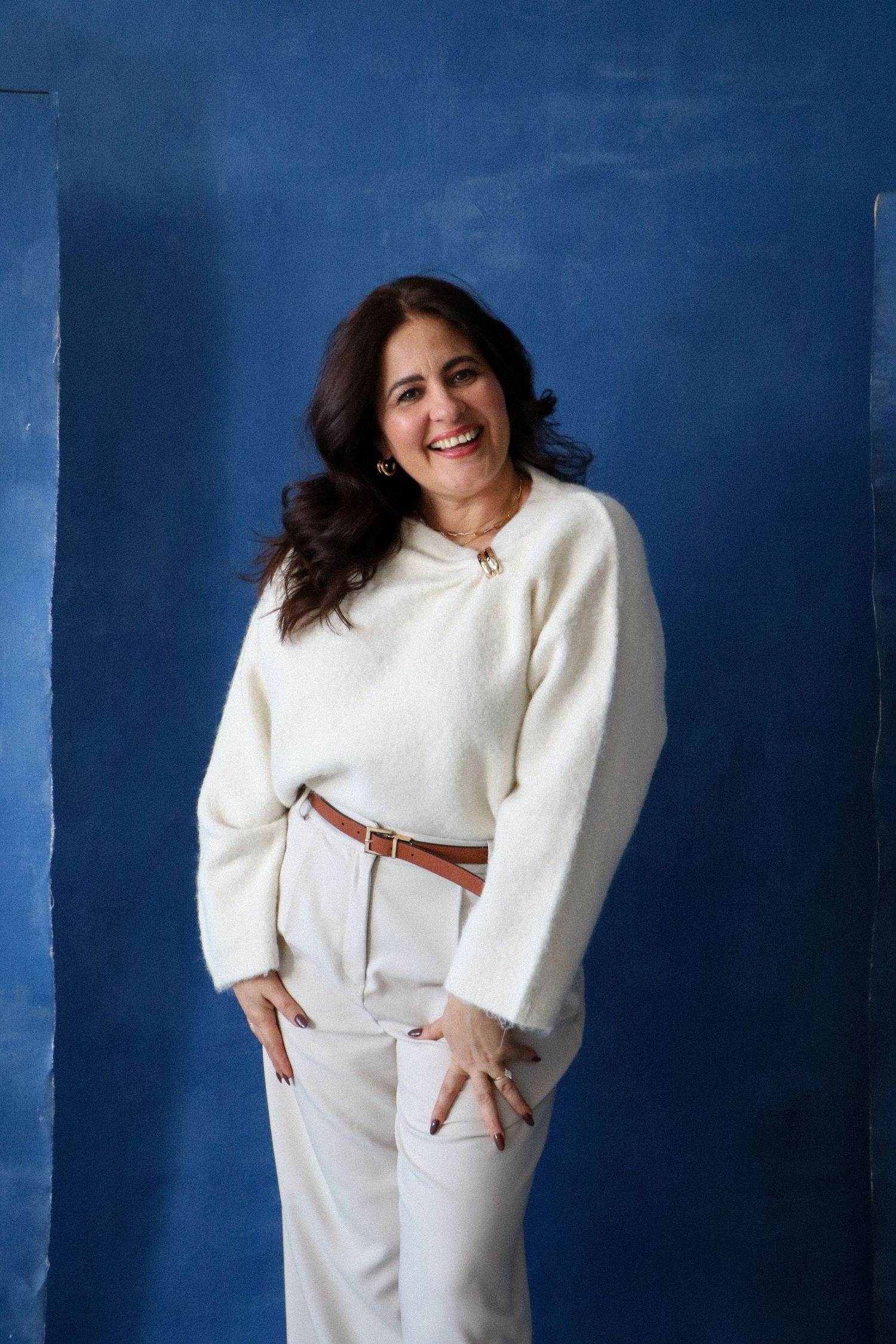 Confident woman in white sweater and pants smiling against a blue background.