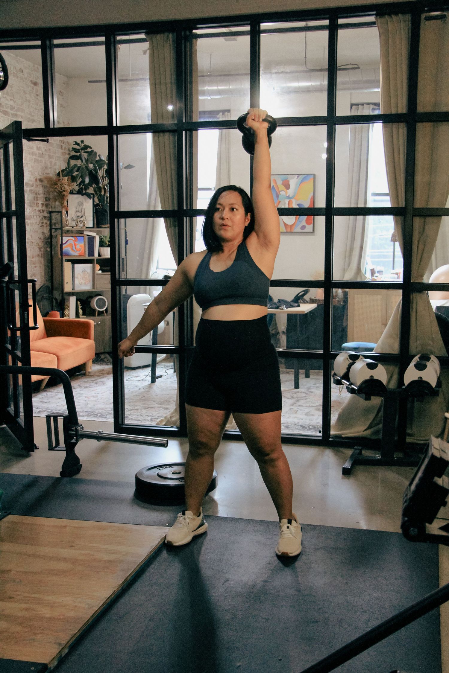 Woman lifting kettlebell overhead in home gym wearing black sports bra and shorts.