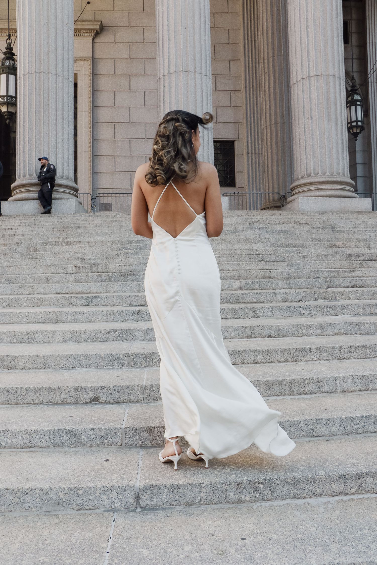 Bride in backless white satin slip gown walks up grand stone steps with tall columns.
