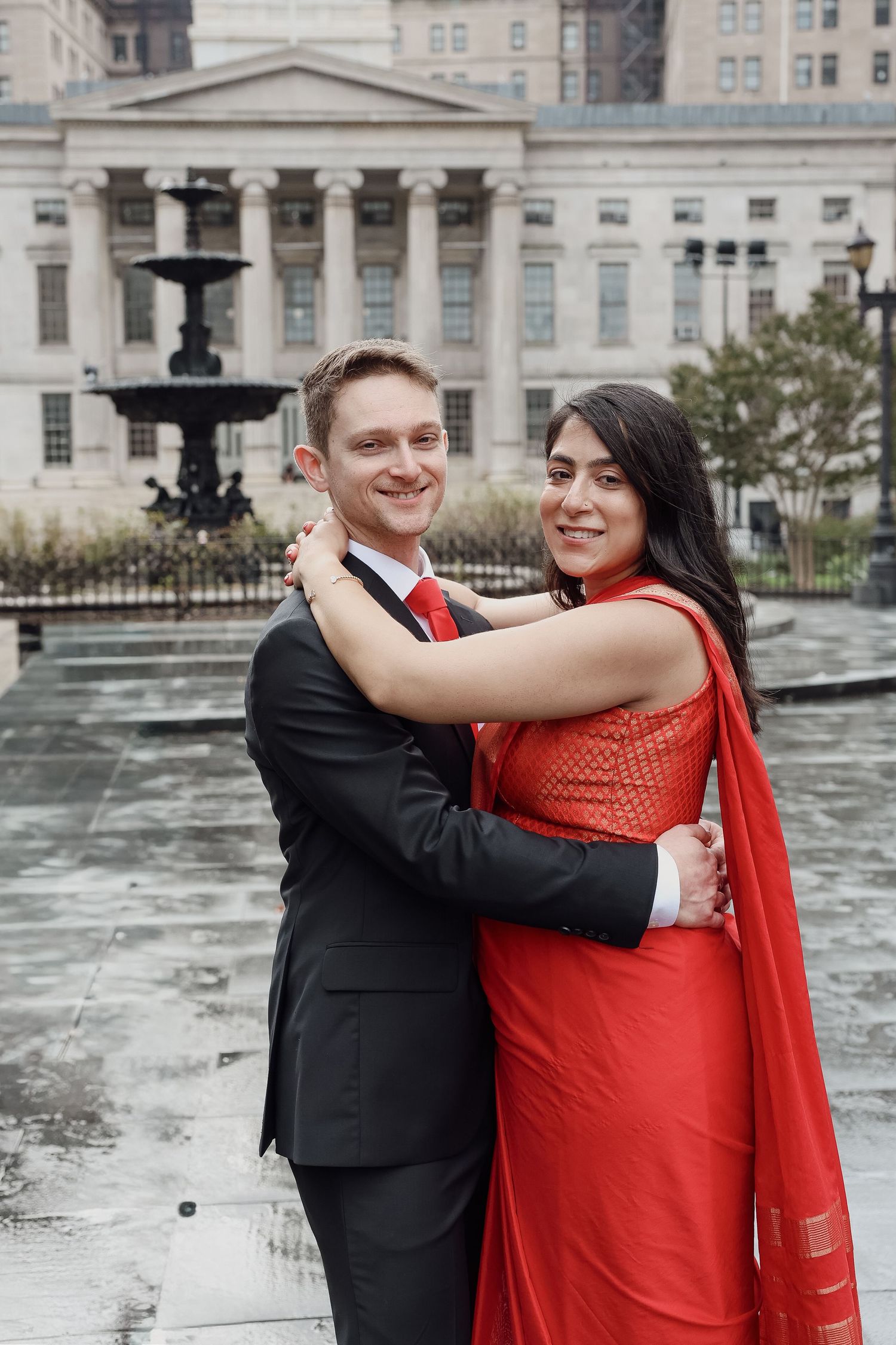 Smiling couple embracing in front of a neoclassical building and fountain on wet pavement, he in a dark suit and red tie, she in a red sari.