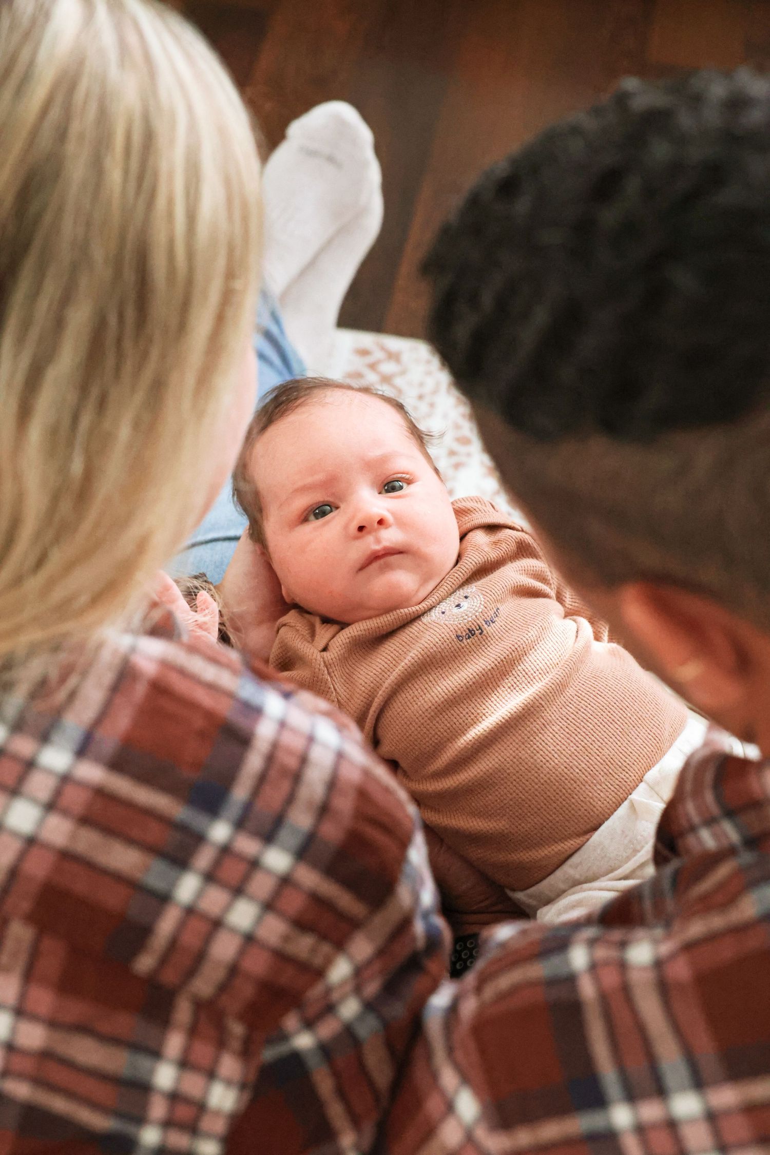 Newborn baby in a tan onesie gazes up at two parents in plaid flannel shirts as they hold and look down at the infant.