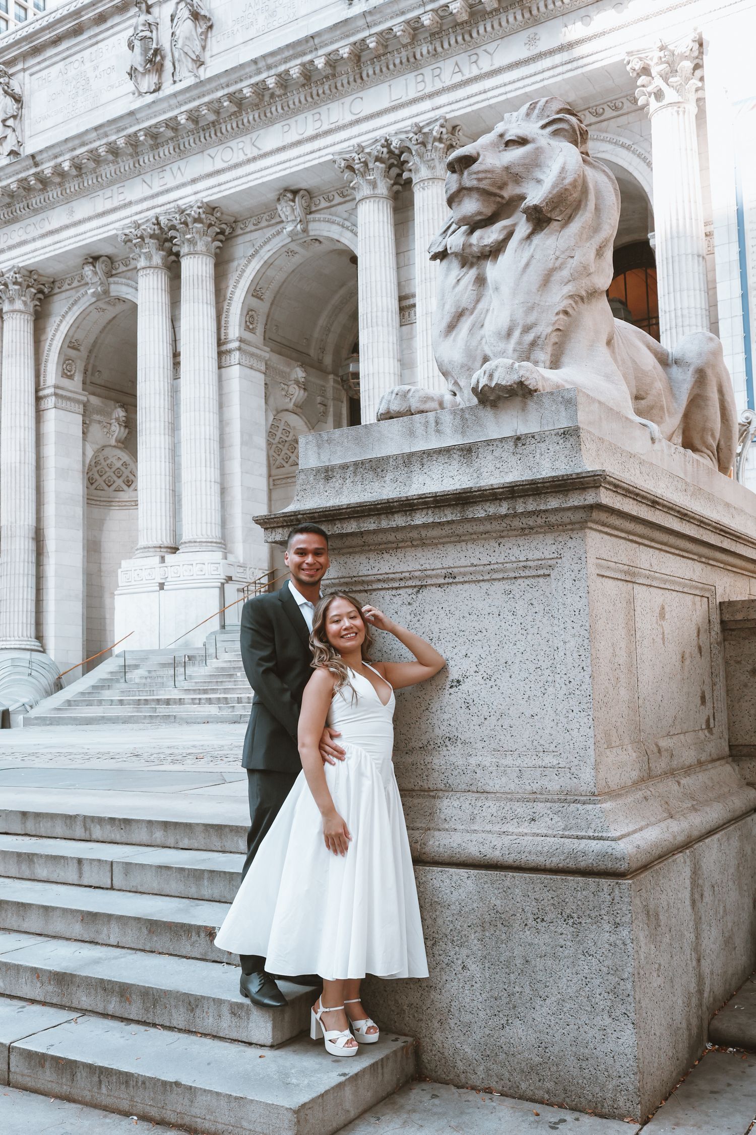 Couple in wedding attire embracing on marble steps beside a large stone lion statue at a grand neoclassical building.