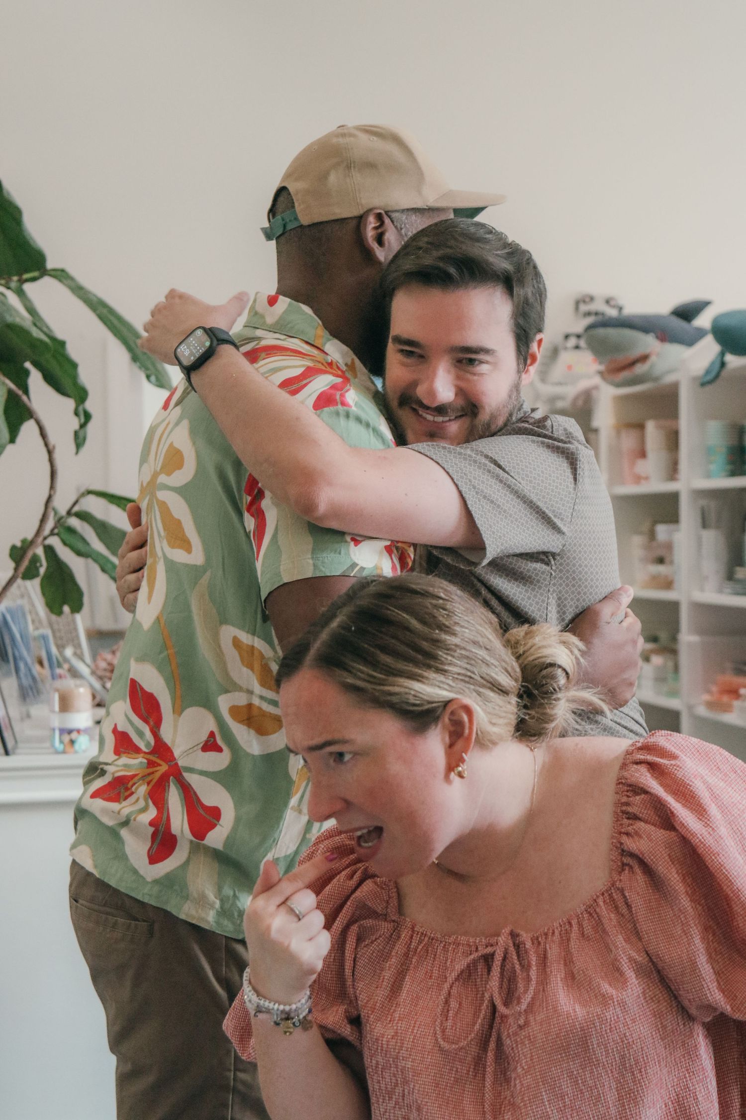 Two men in a warm hug in a bright shop while a woman in a pink blouse reacts excitedly in the foreground.