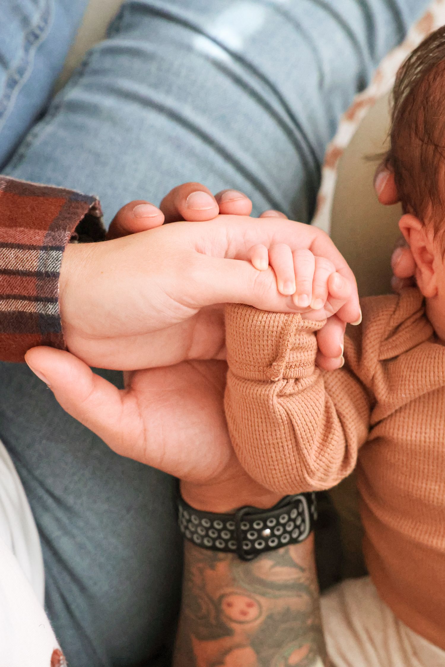 Tattooed adult hands gently hold a newborn's tiny hand, with the baby in a tan knit outfit resting against a plaid sleeve and jeans.