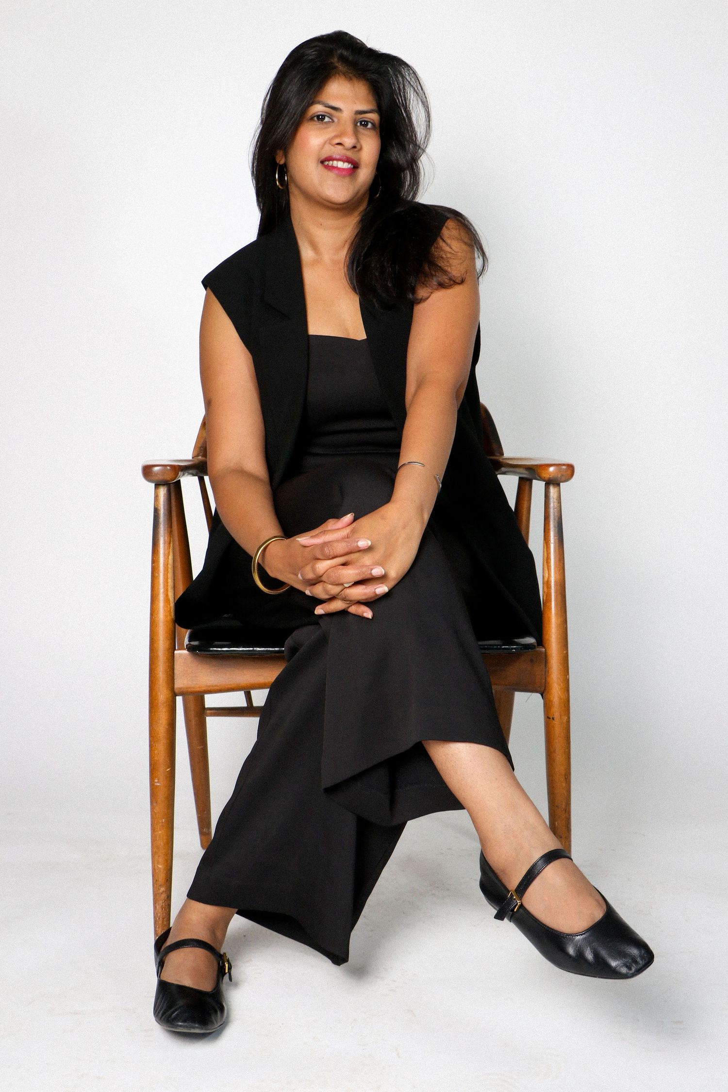 Confident woman in all-black outfit sitting relaxed in a wooden chair against a clean white background.