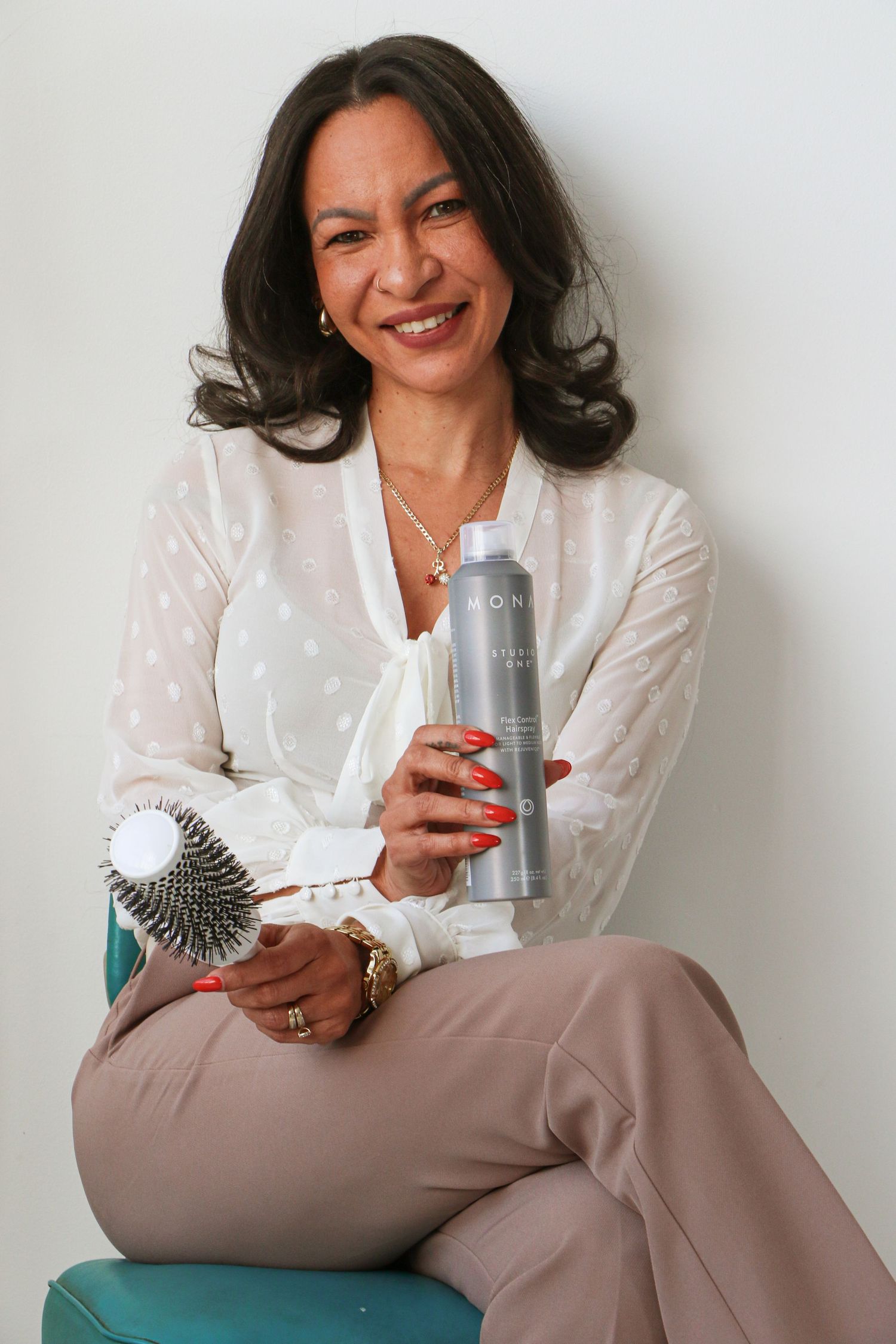 Smiling woman in white blouse holding MONAT hairspray and round brush on teal stool.