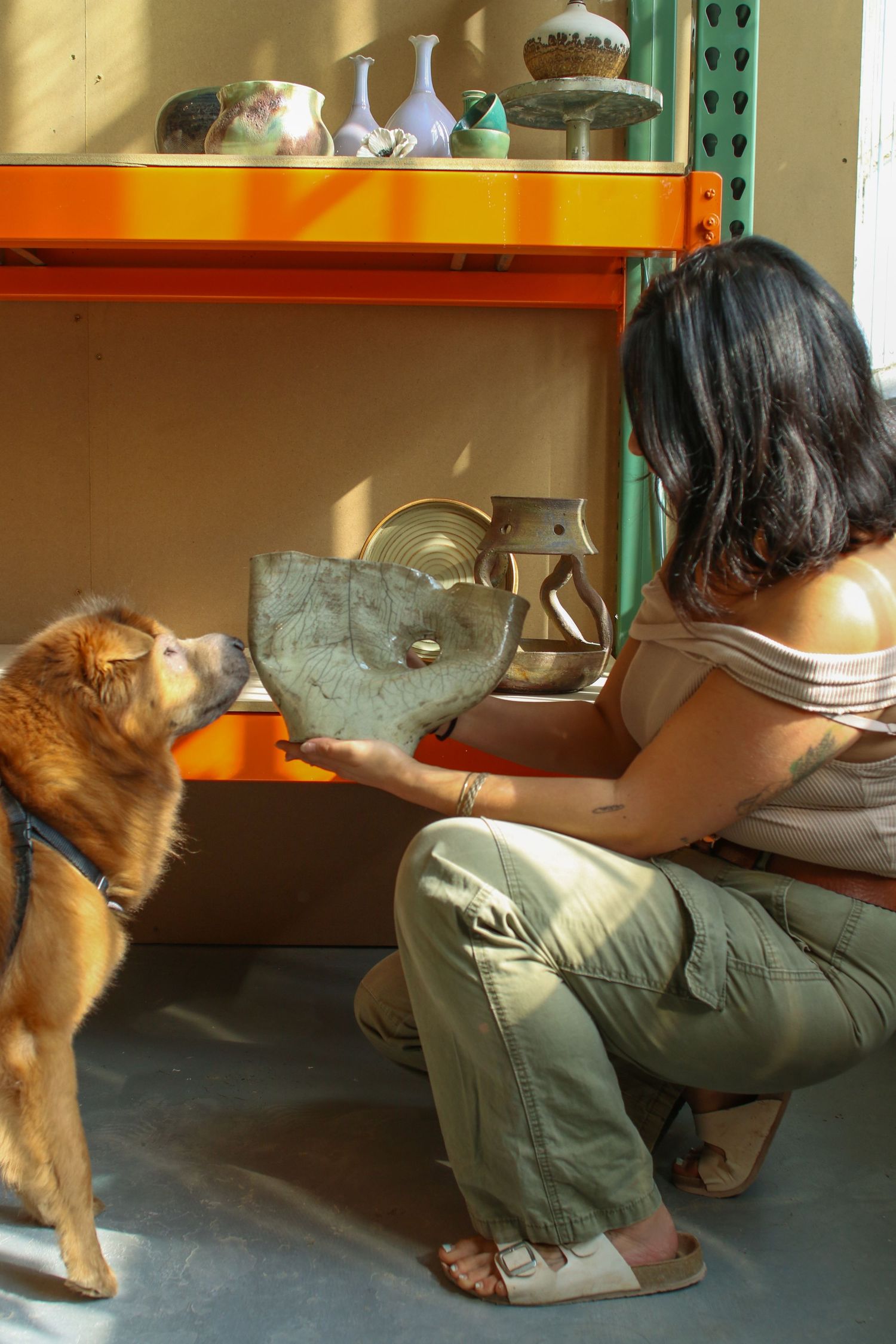 Woman crouching and showing a metal heart sculpture to a brown dog in a pottery studio with ceramic pieces on shelves.