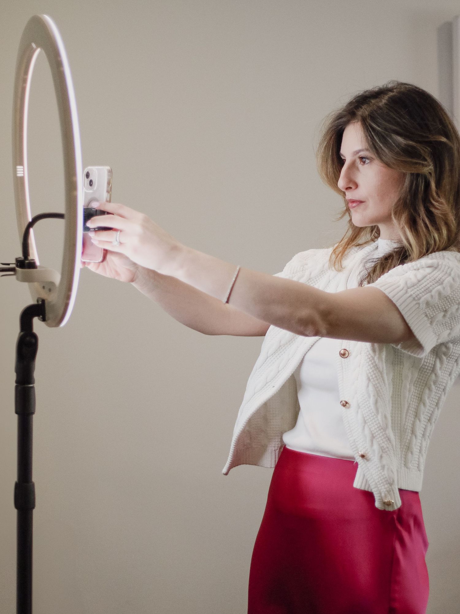 Woman in white knit top and red skirt setting up phone on ring light.