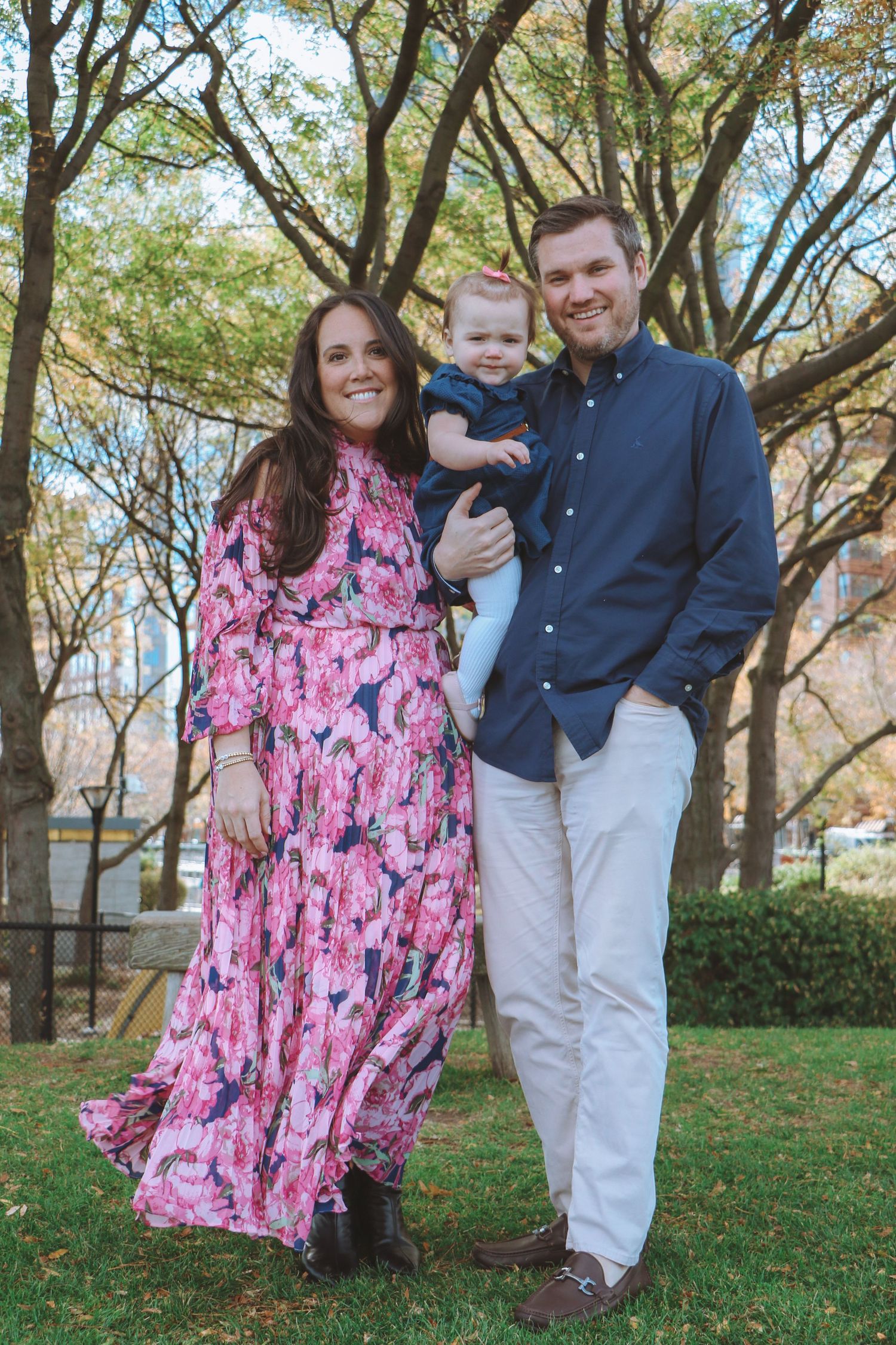 Happy family with baby posing on green grass surrounded by autumn trees in urban park.