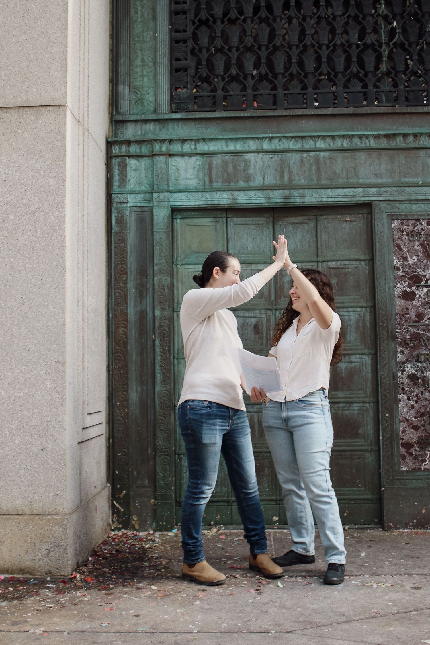 Two women high-fiving joyfully outside a building with ornate green bronze doors.