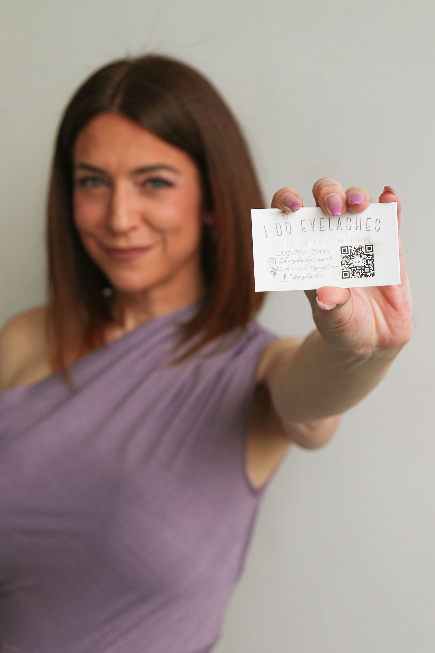 Woman in purple dress holding a business card for I Do Eyelashes with a QR code toward the camera.