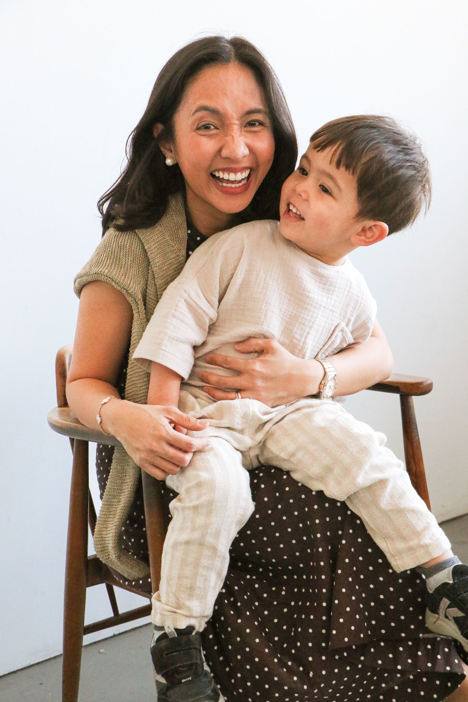 Asian mother laughing joyfully with young son on her lap in wooden chair, white background.