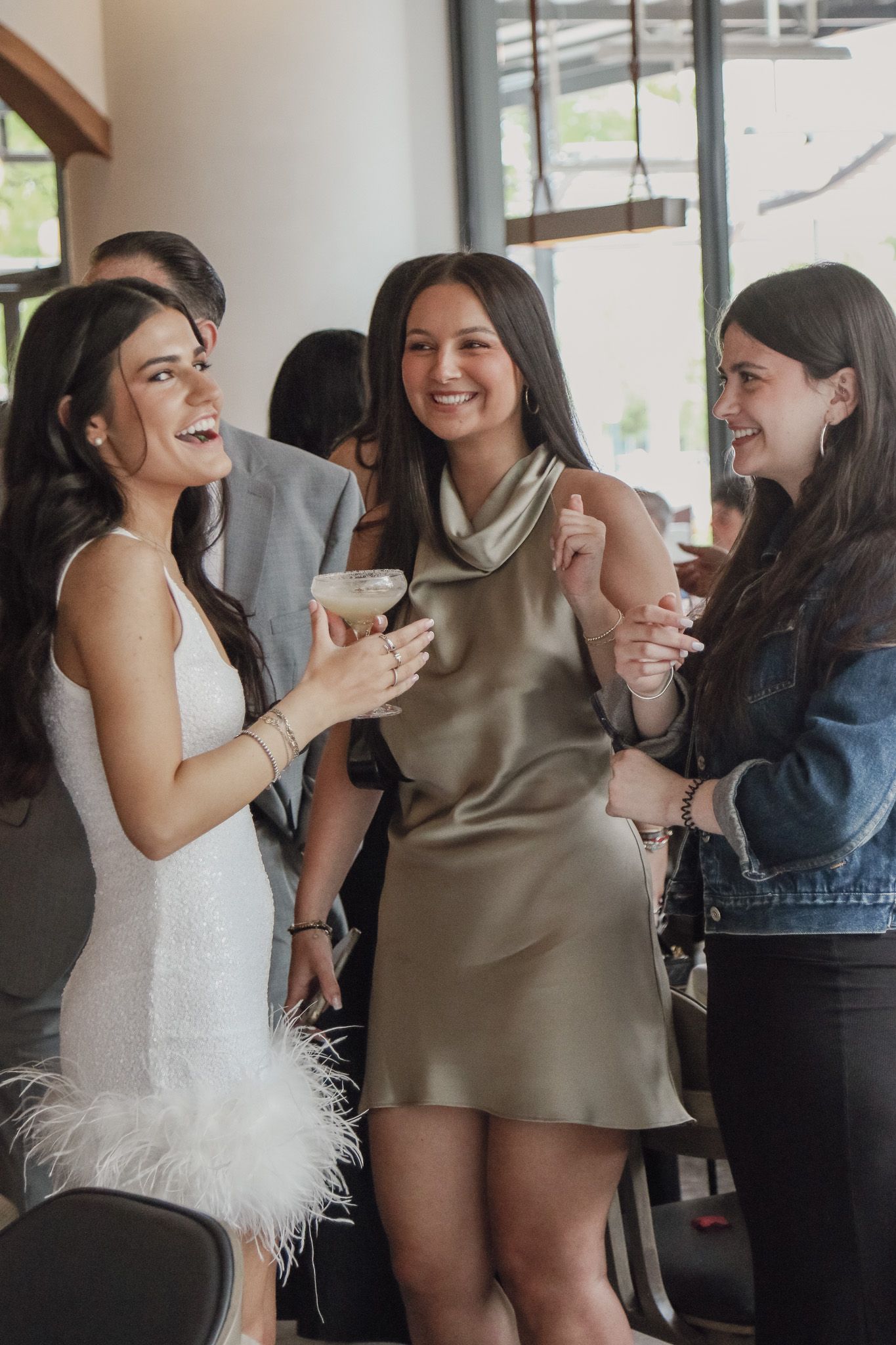 Three smiling women in stylish dresses and a denim jacket laugh together while holding drinks at a lively indoor gathering.