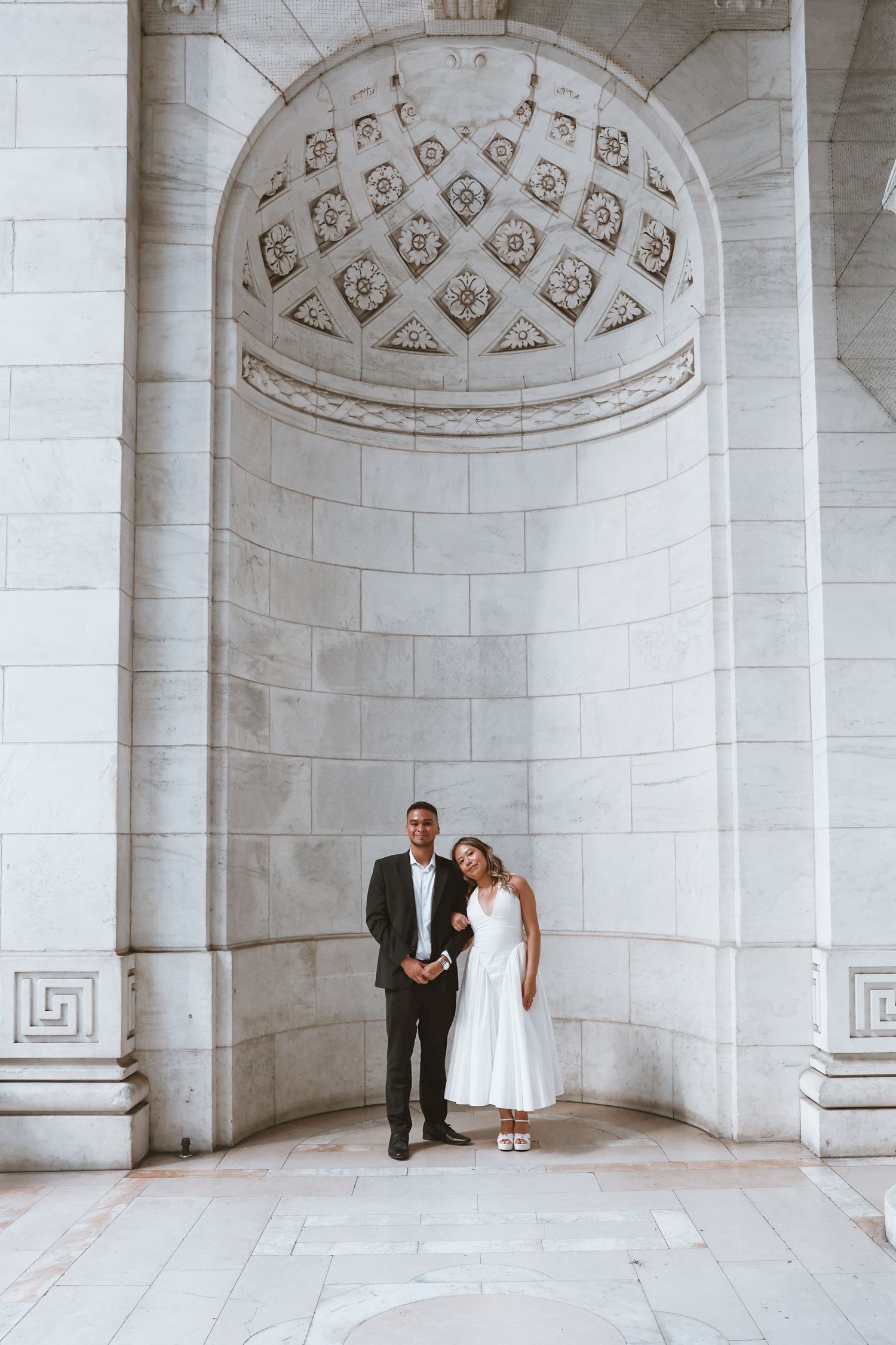 Couple in wedding attire embracing beneath an ornate coffered marble alcove with carved stone details and a geometric Greek key pattern.