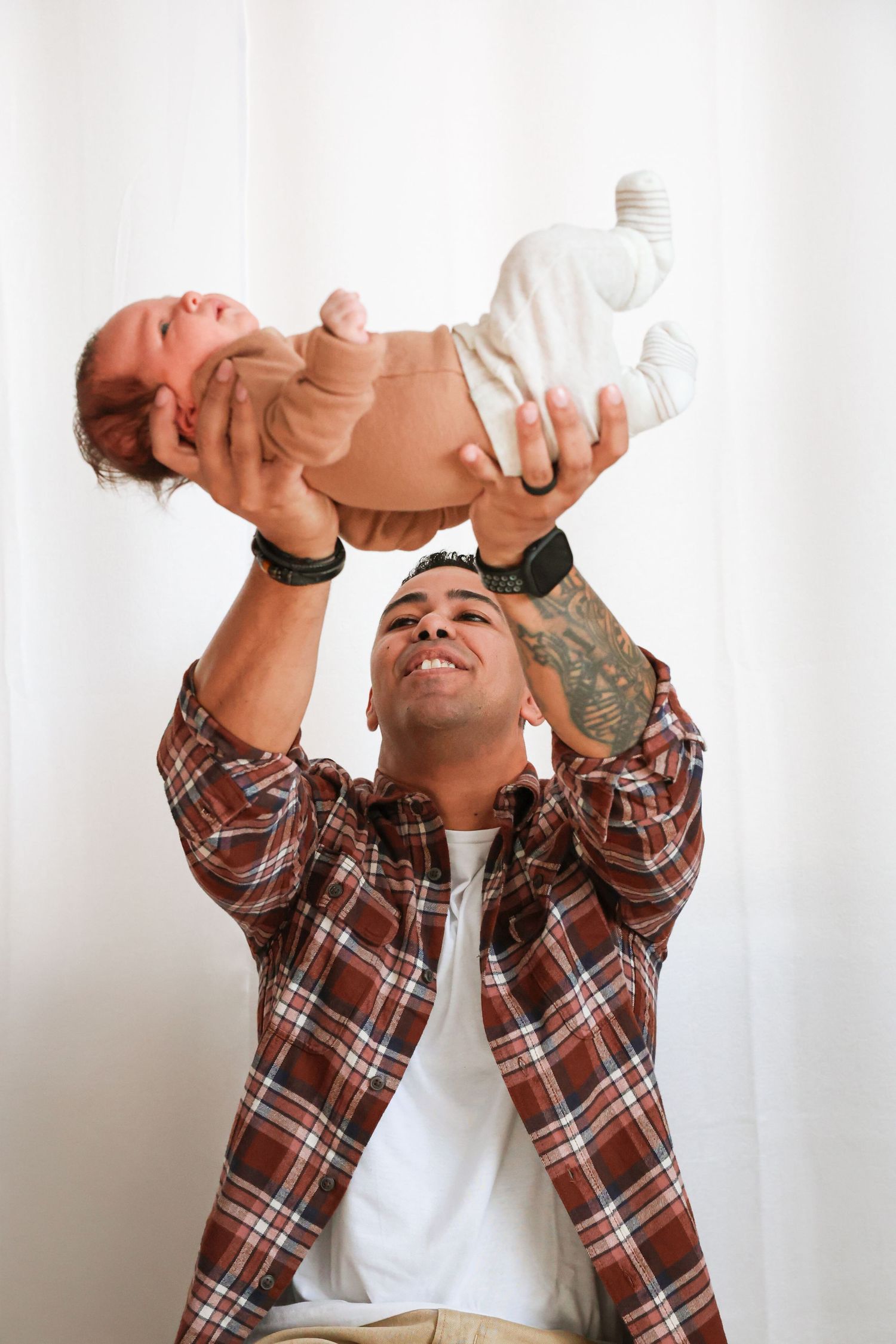 Smiling tattooed father in a plaid flannel shirt lifts his sleeping newborn baby overhead against a soft white curtain backdrop.