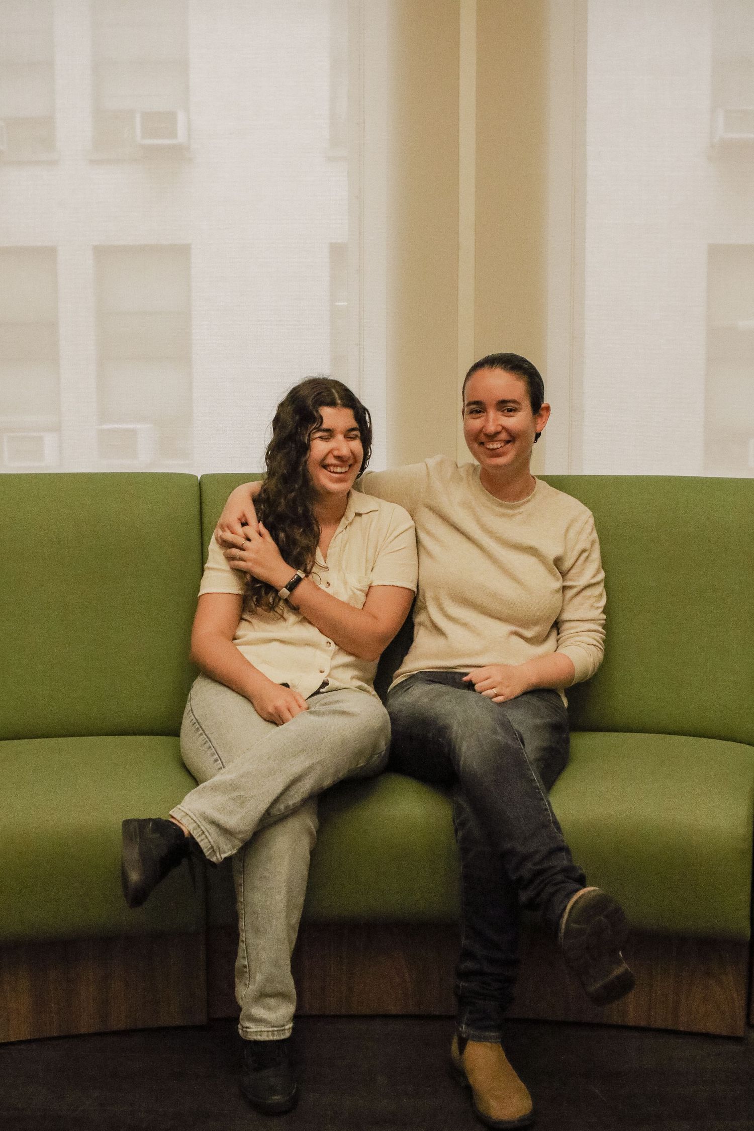 Two women smiling and posing together on a green curved sofa near bright windows.