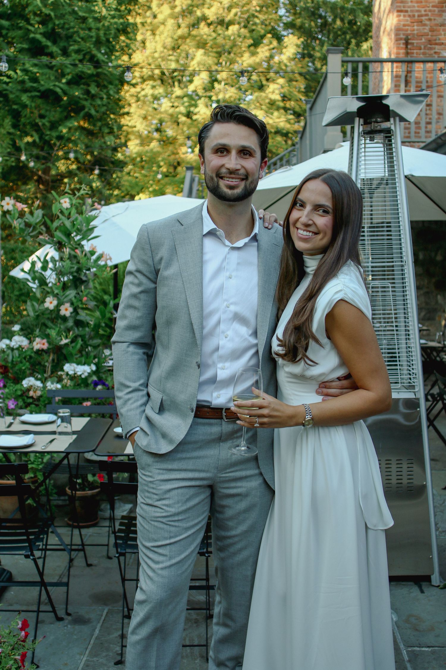 Smiling couple in elegant attire posing together on a garden patio, man in grey suit, woman in white dress.