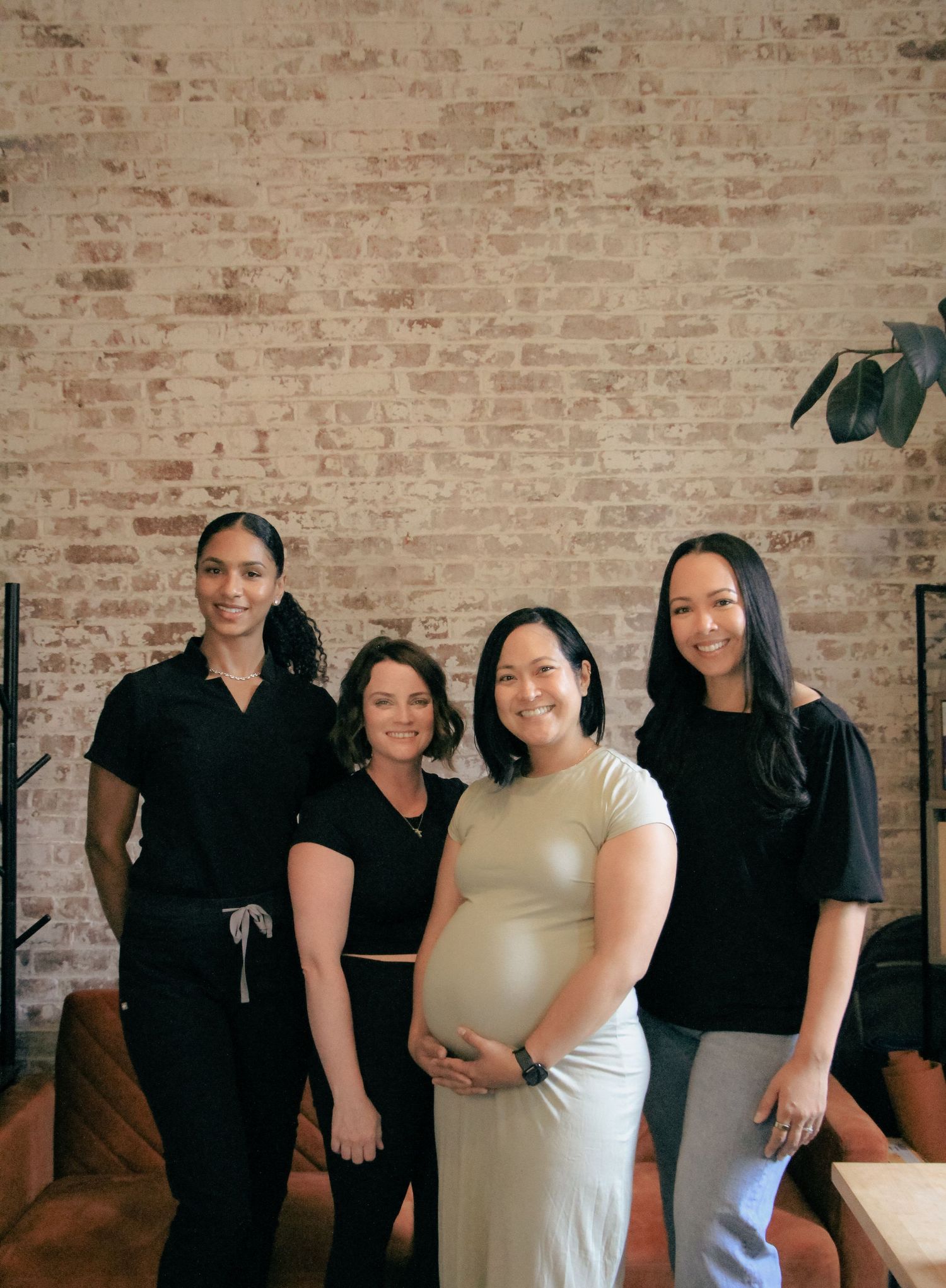 Four women posing together in front of exposed brick wall, one visibly pregnant holding her baby bump.