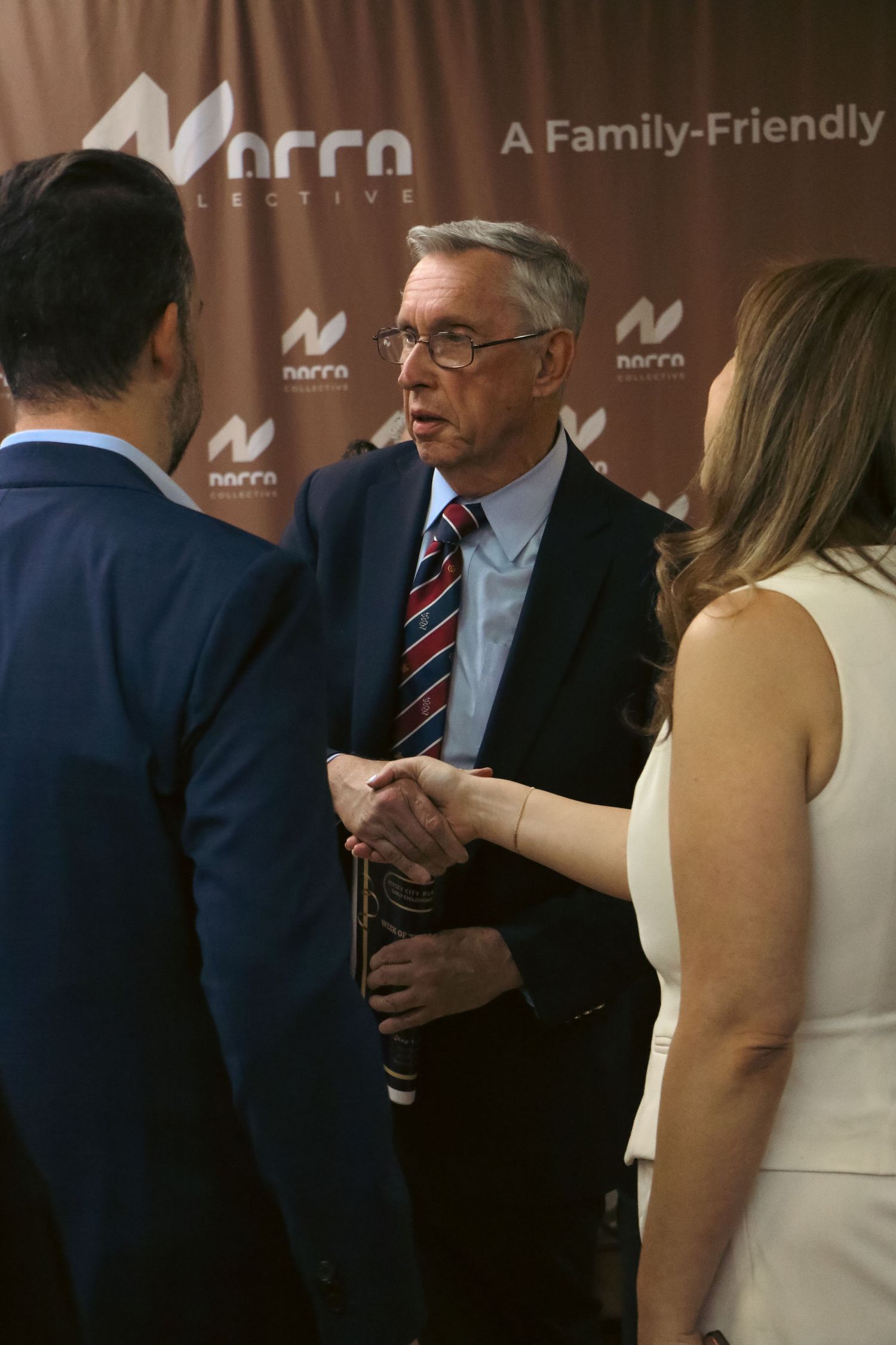 Man in suit shaking hands at a Norra Collective family-friendly event backdrop.
