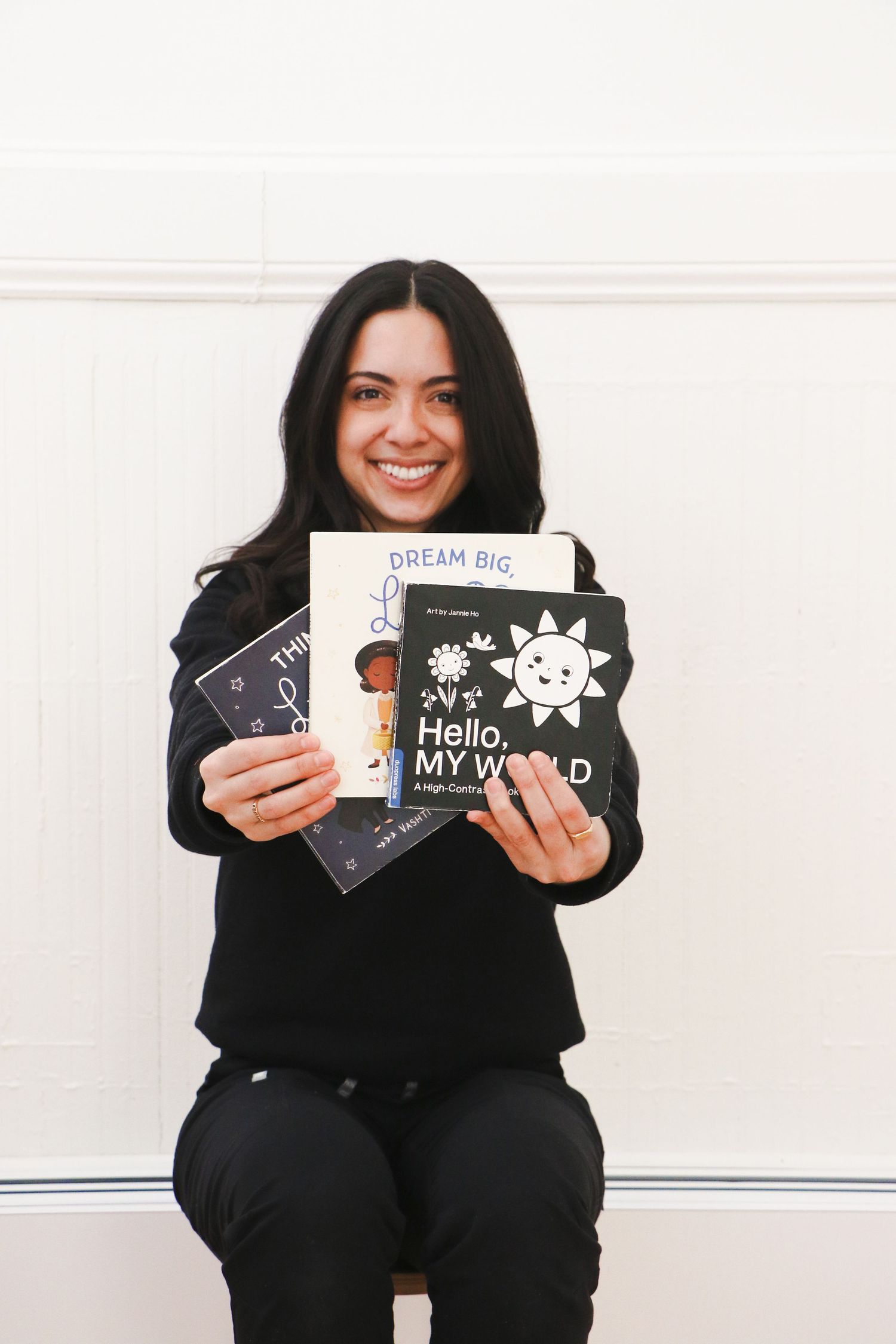 Smiling young woman in black outfit holding children's board books including Hello My World while seated.