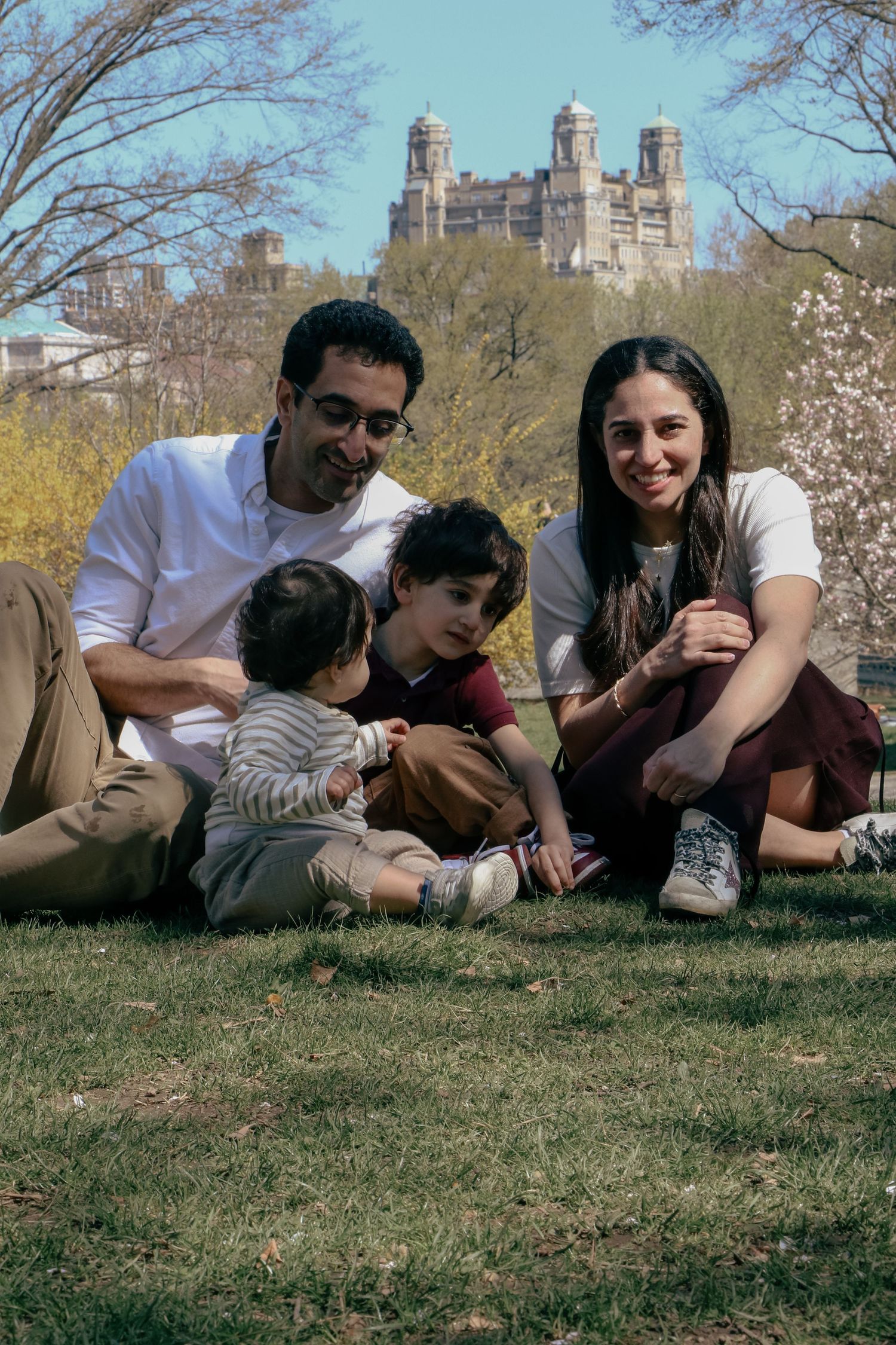 Happy family of four sitting on grass in Central Park with historic building visible in background.
