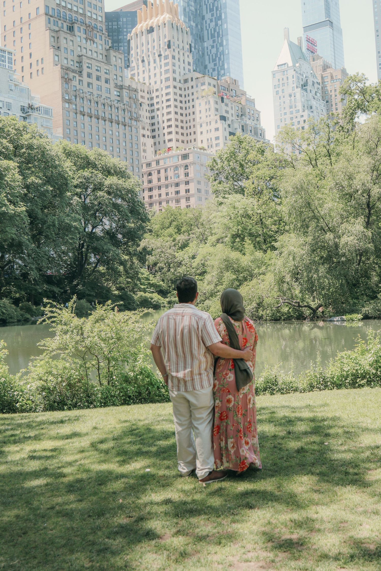 Couple standing arm in arm on a sunny grassy lawn, gazing at a calm pond with lush trees and tall city skyscrapers in the distance.
