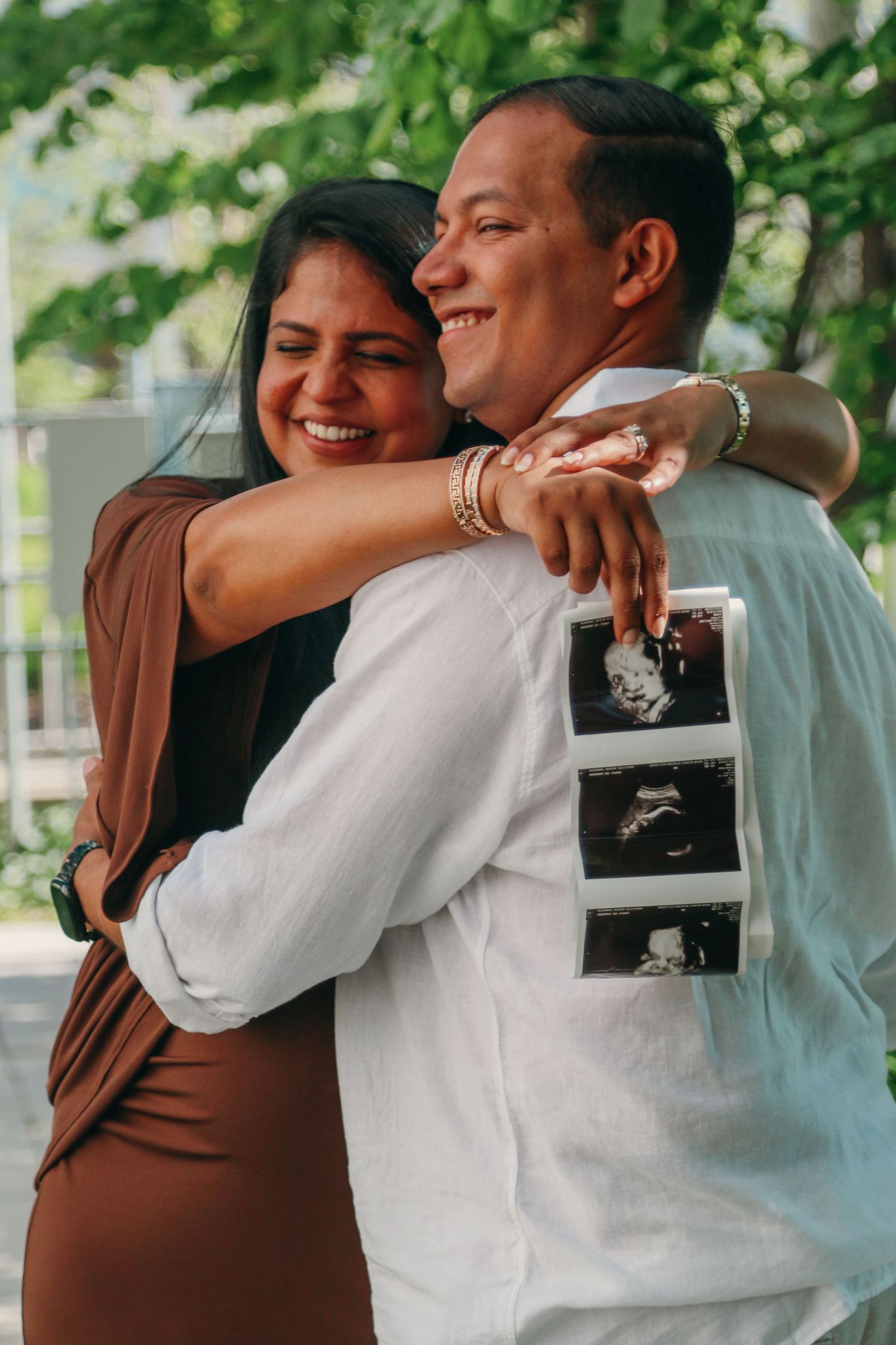 Happy couple hugging outdoors while holding ultrasound photos, announcing pregnancy with joyful smiles.