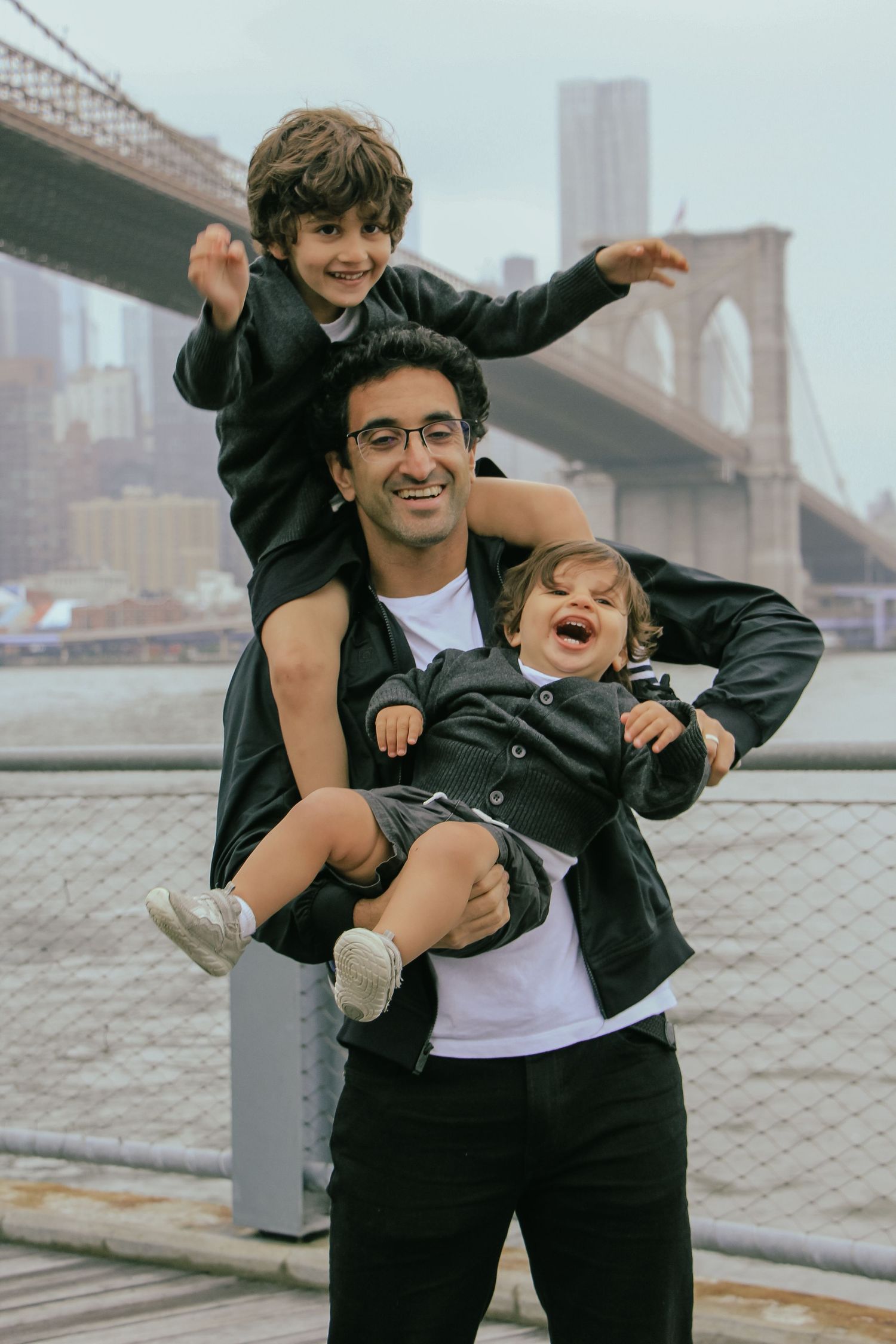 Smiling dad holding two young children near Brooklyn Bridge waterfront.
