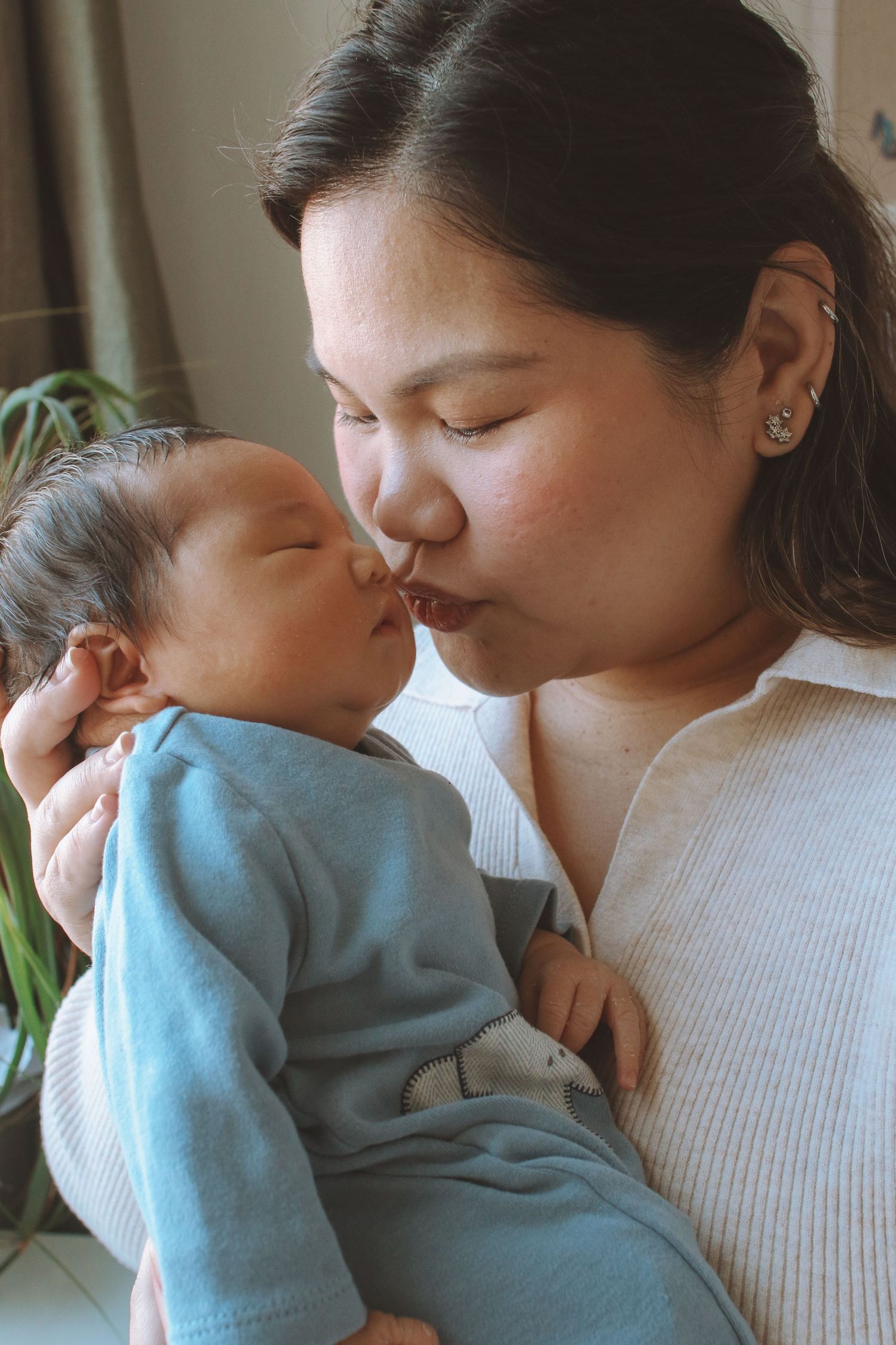Asian mother gently kissing her newborn baby dressed in blue onesie.