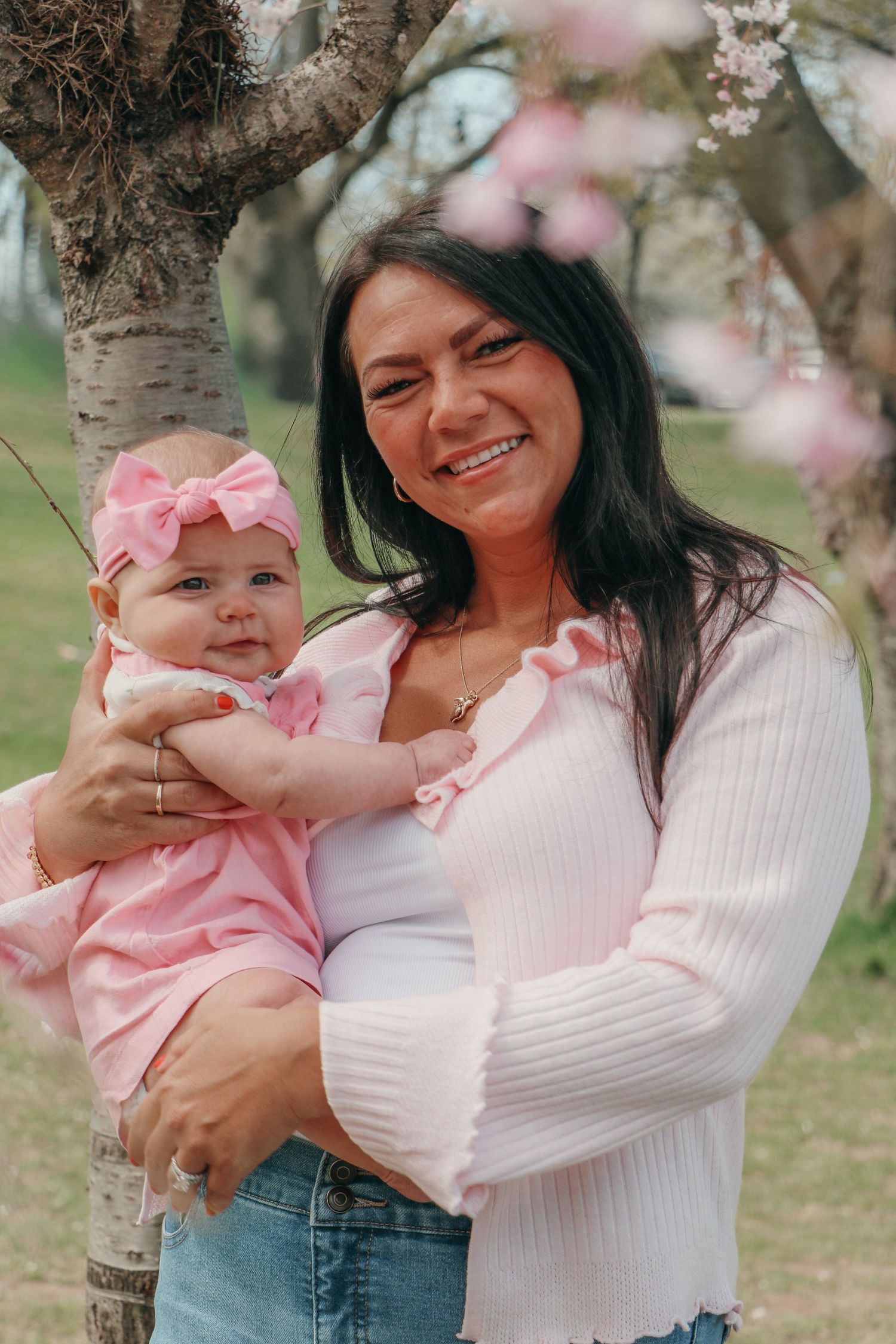 Smiling dark-haired mother holding baby girl in pink dress under blooming cherry blossom tree.