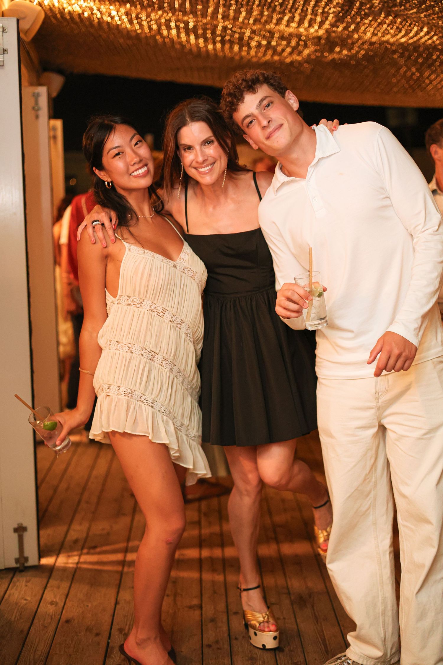 Two smiling women in white and black dresses pose with a young man in white clothes, holding drinks at an evening party on a wooden deck.