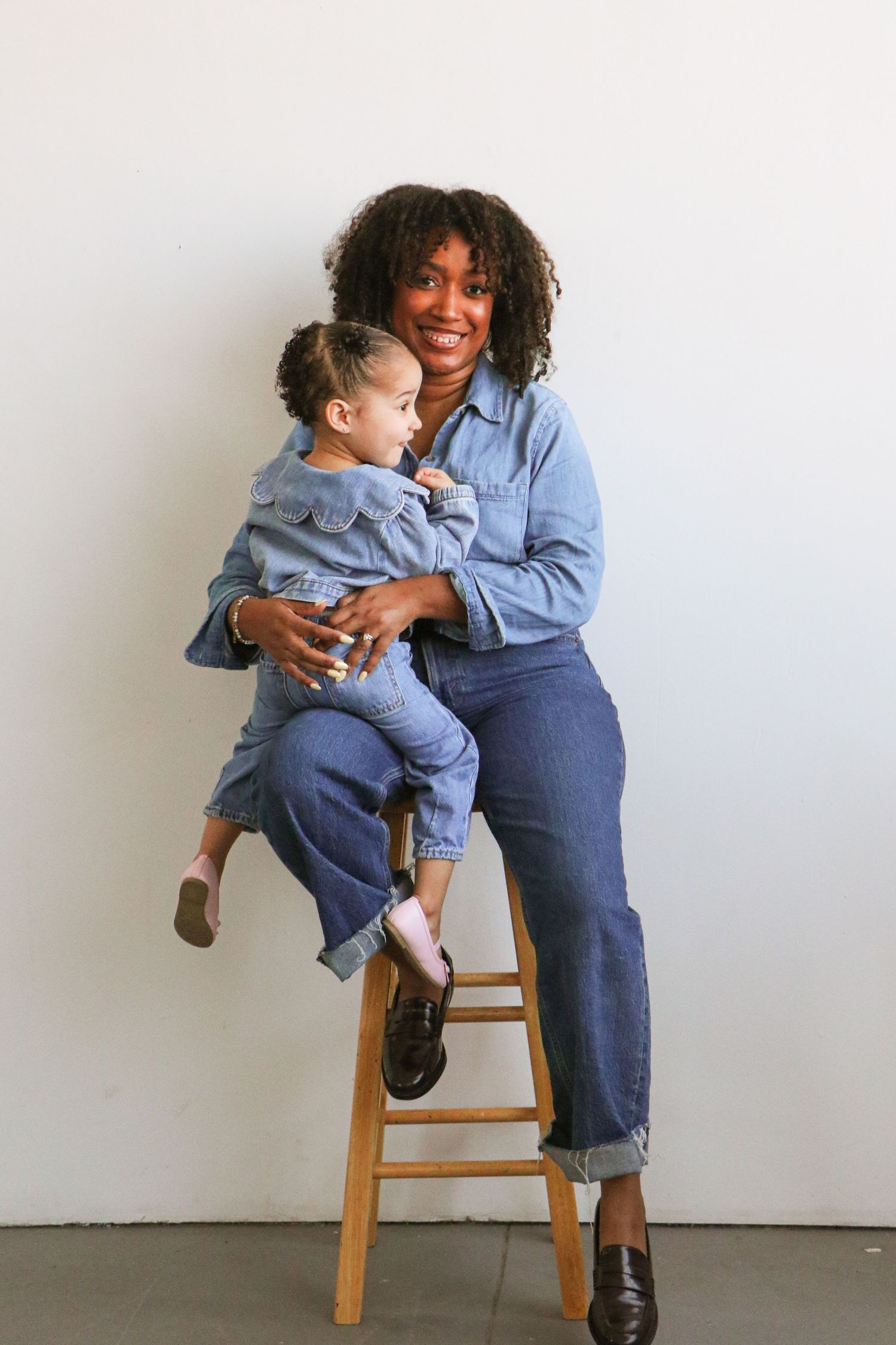 Smiling Black mother in denim sitting on stool holding toddler daughter in matching denim.