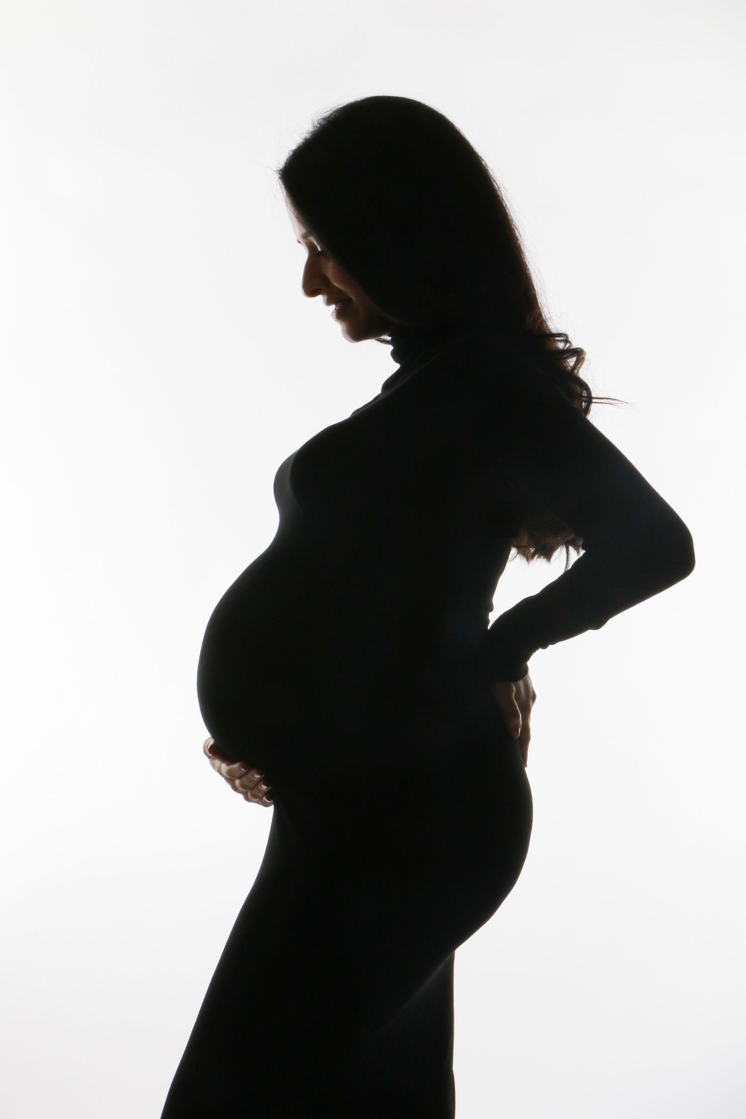 Silhouette of pregnant woman cradling baby bump against bright white background.