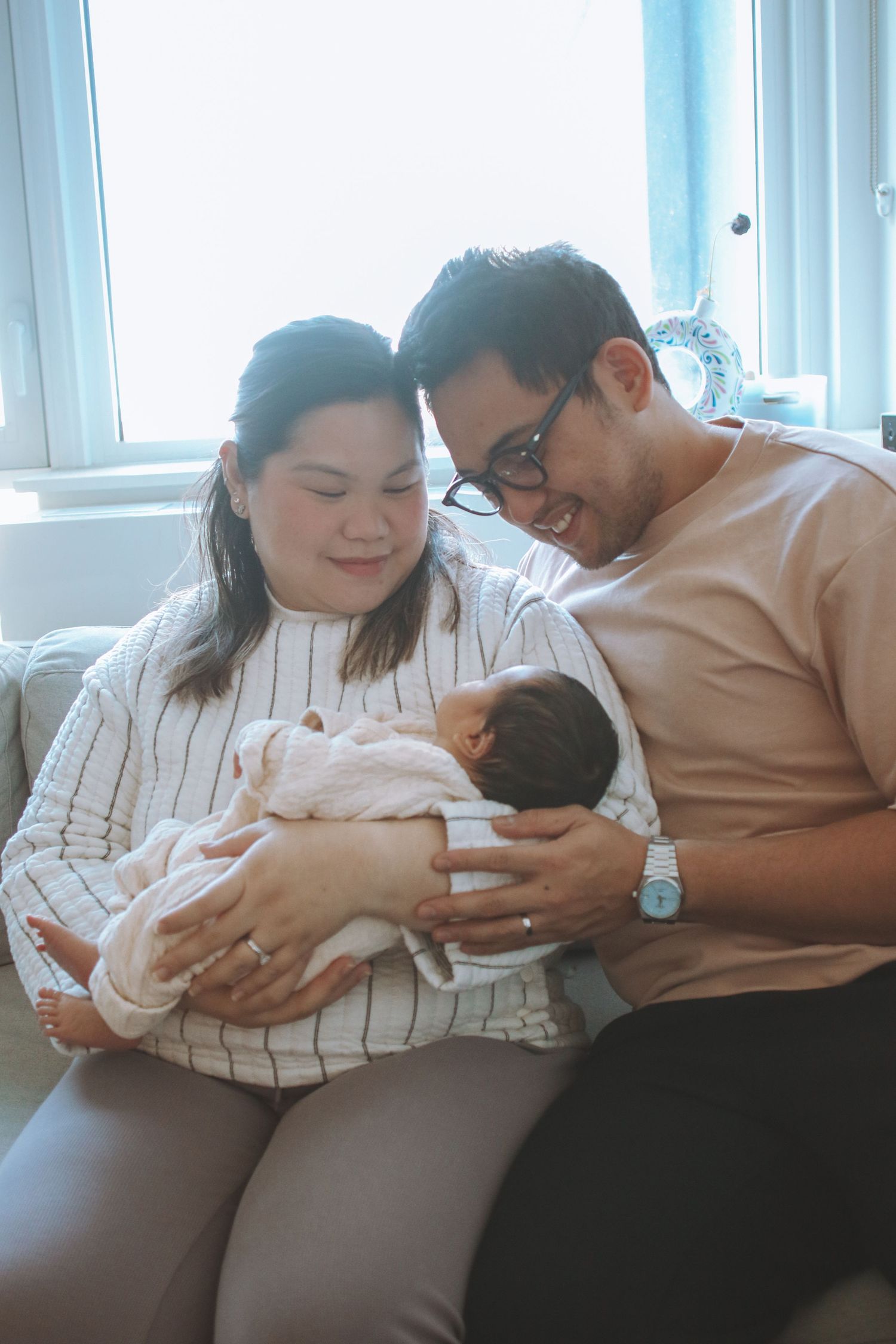 Asian couple sitting on couch smiling at their newborn baby wrapped in white blanket.