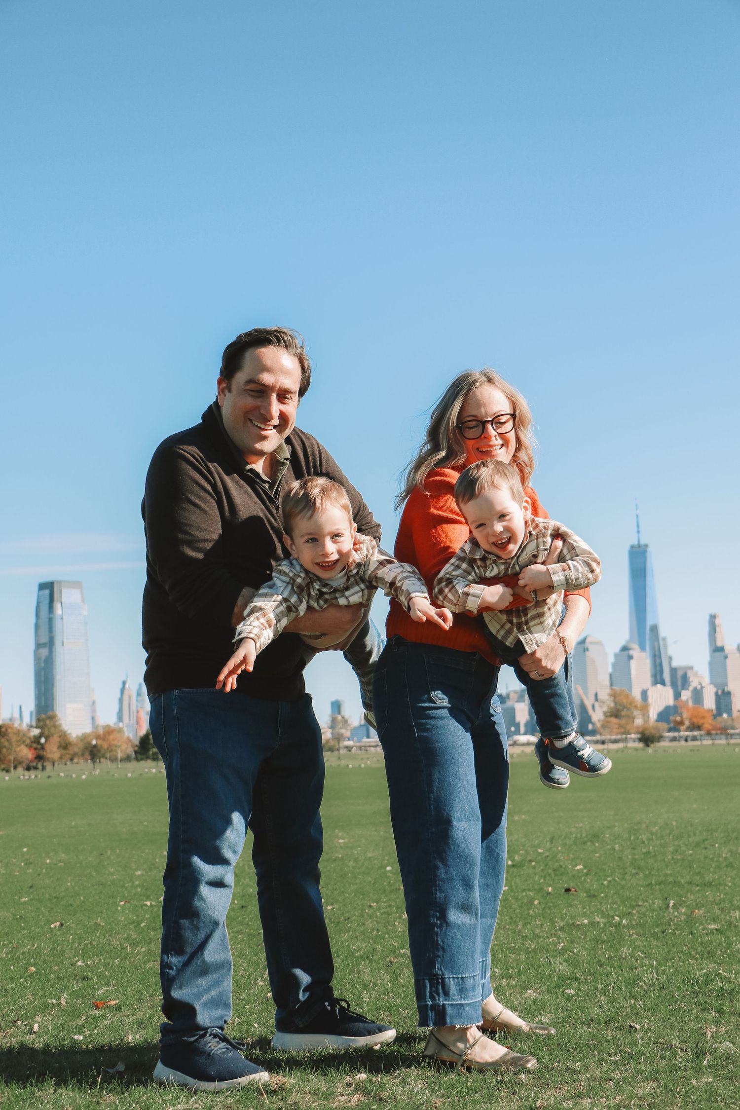 Family of four with twins posing on green field with NYC skyline backdrop on a sunny day.