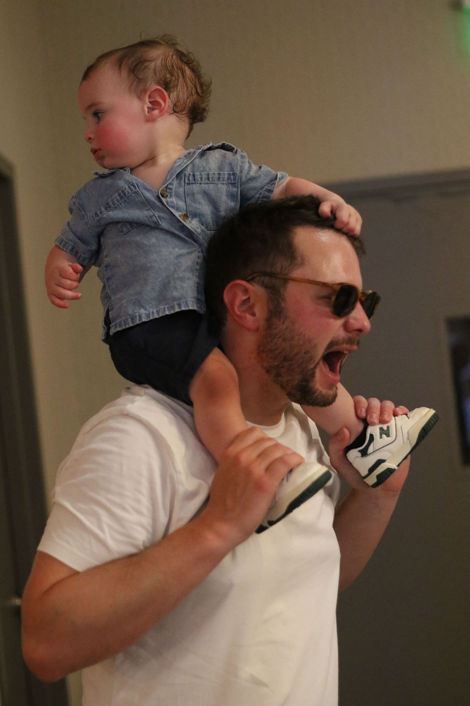 Laughing man in sunglasses carrying a baby in a denim jacket on his shoulders indoors.