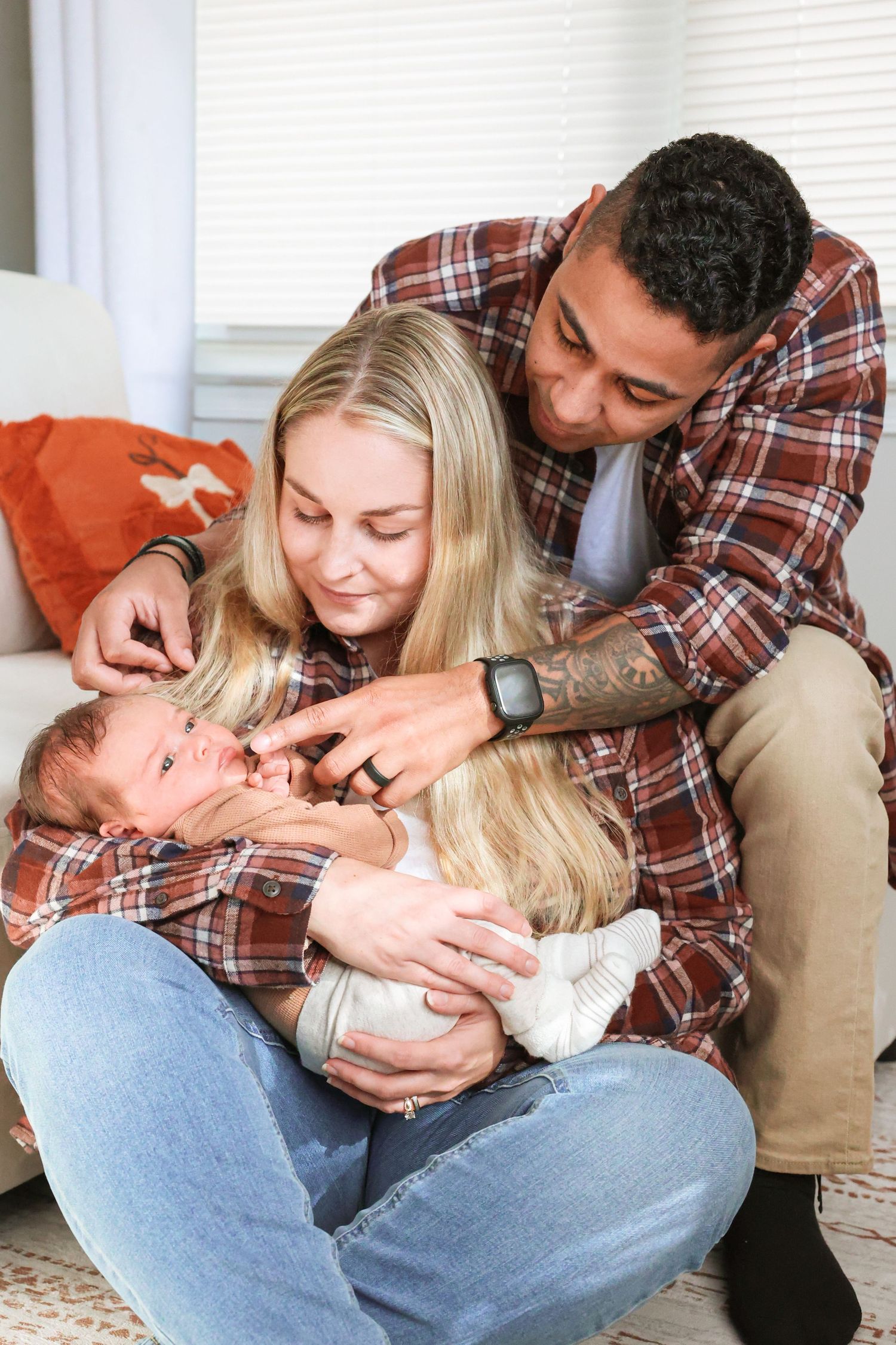 Smiling couple in matching plaid flannel shirts sit on the floor gently cradling and touching their newborn baby in a bright living room.