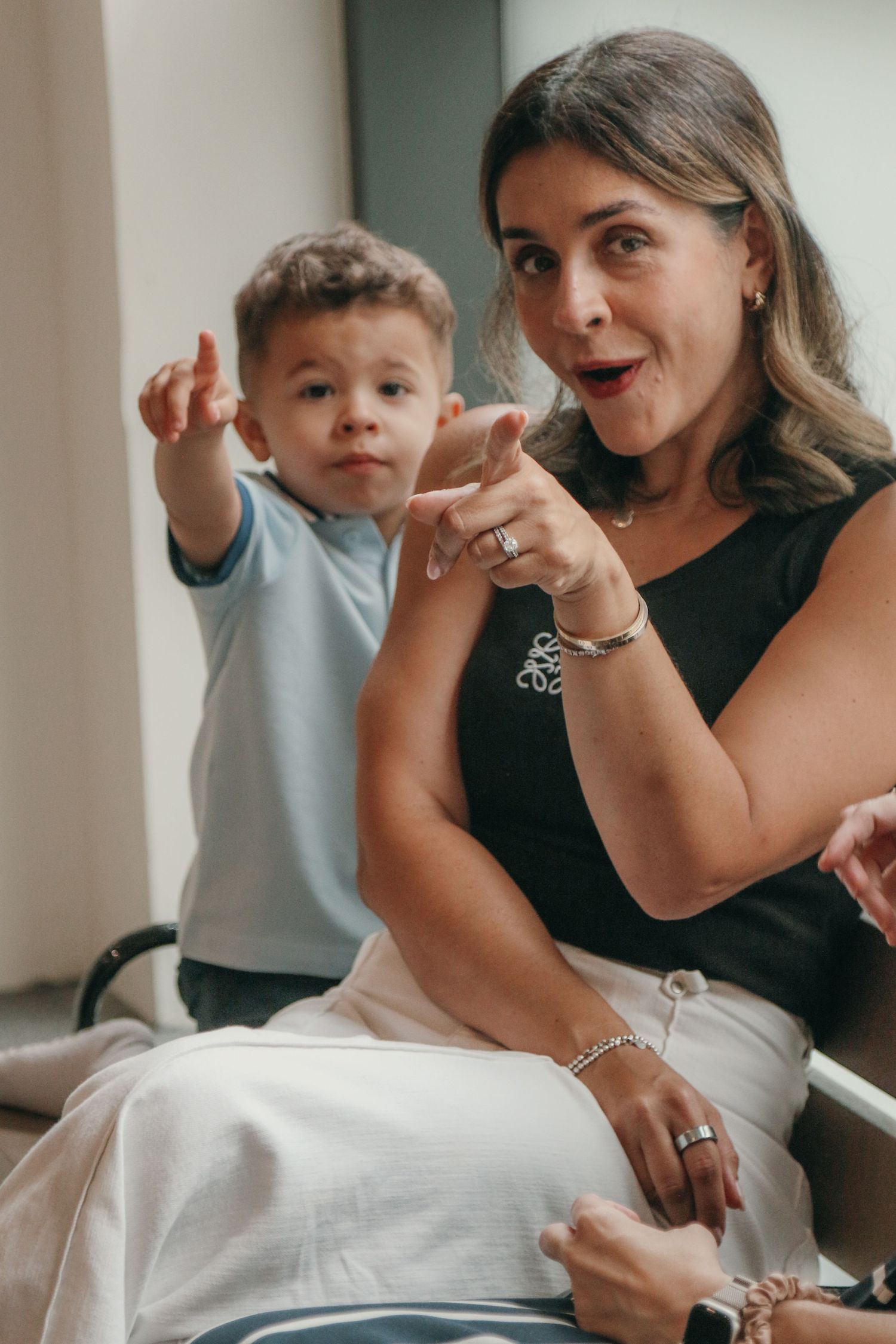 A smiling woman in a black top and white skirt sits beside a toddler in a light blue shirt as both point playfully toward the camera.