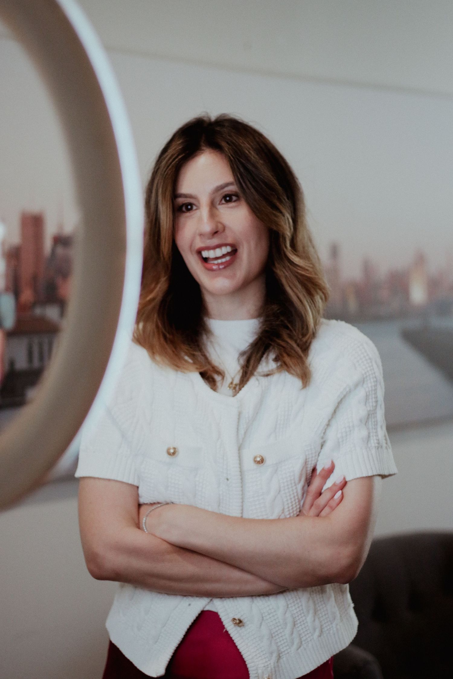 Confident woman in white knit vest with arms crossed smiling in bright room.