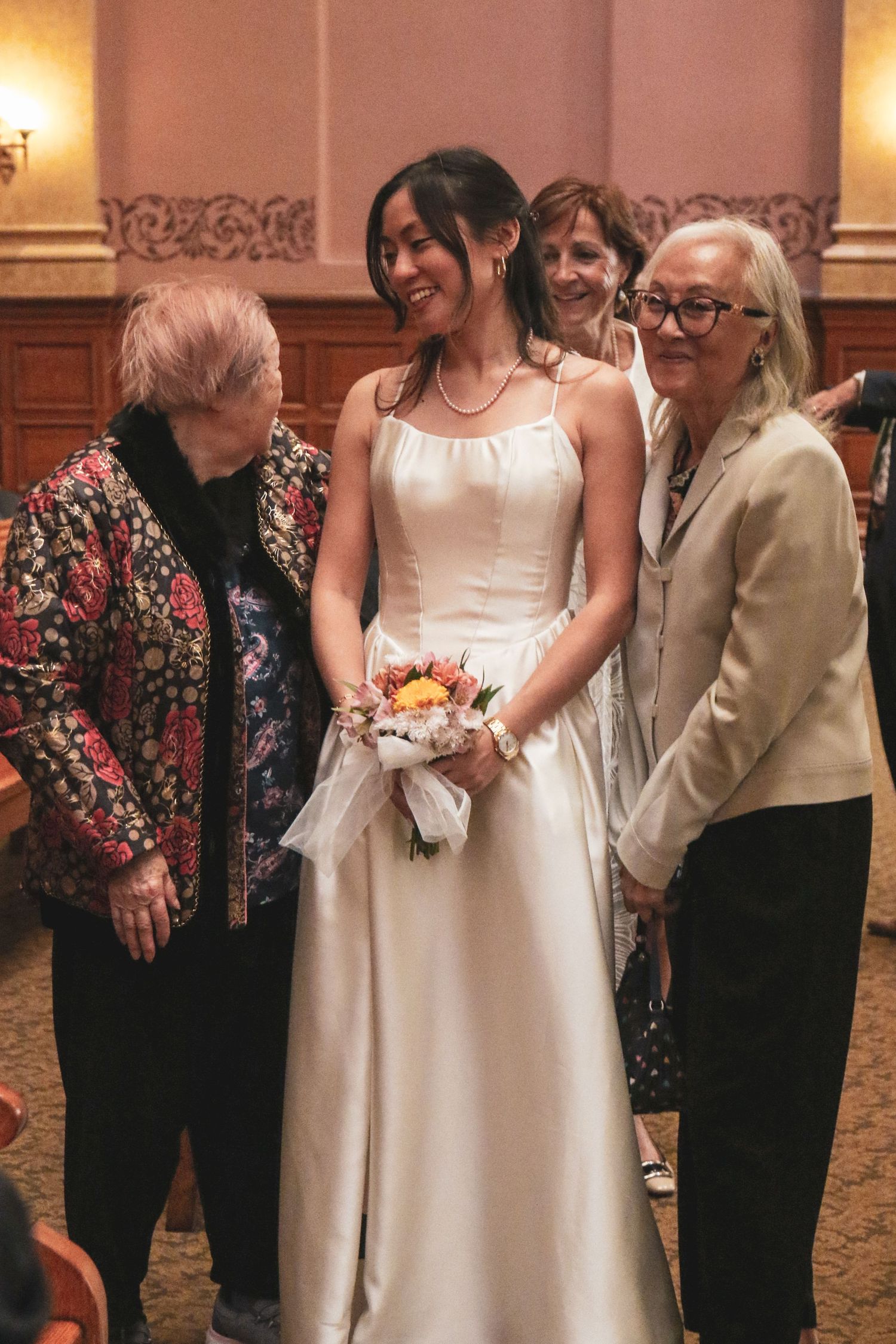 Smiling bride in white gown holding colorful bouquet surrounded by guests at elegant wedding venue.