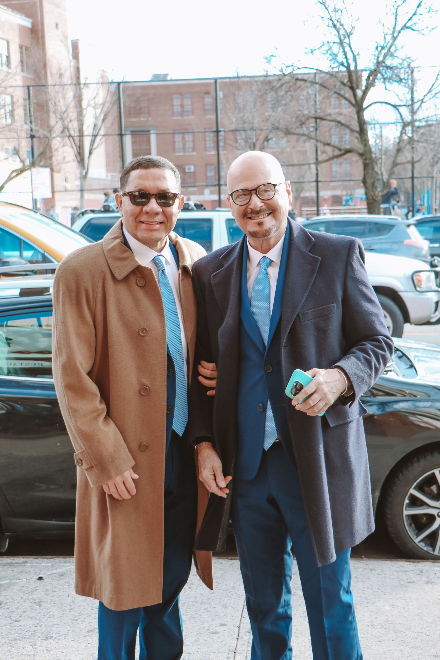 Two well-dressed men in blue suits and ties smiling together outdoors in an urban setting.