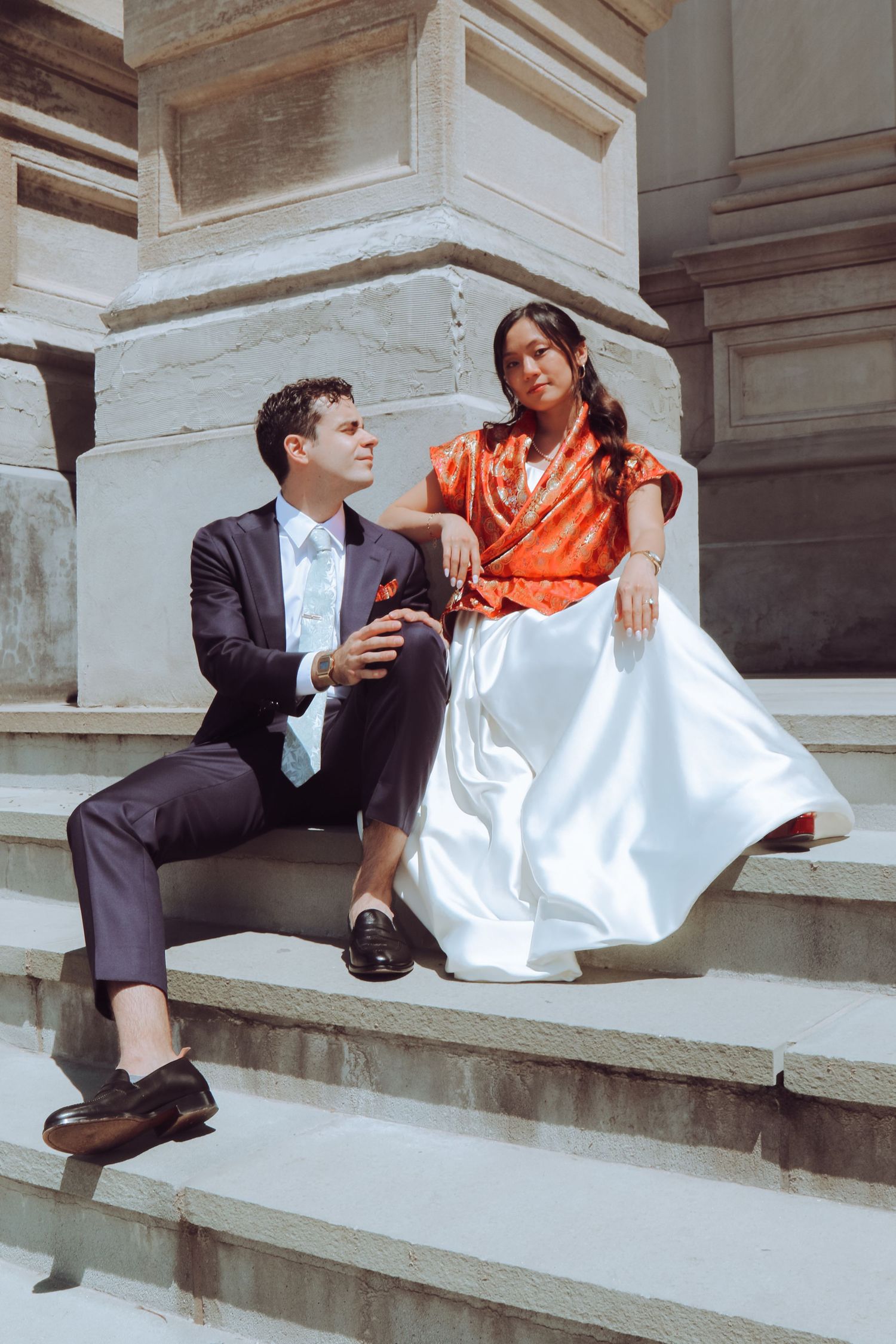 Couple in elegant attire sitting on stone steps, man in dark suit, woman in orange top and white skirt.