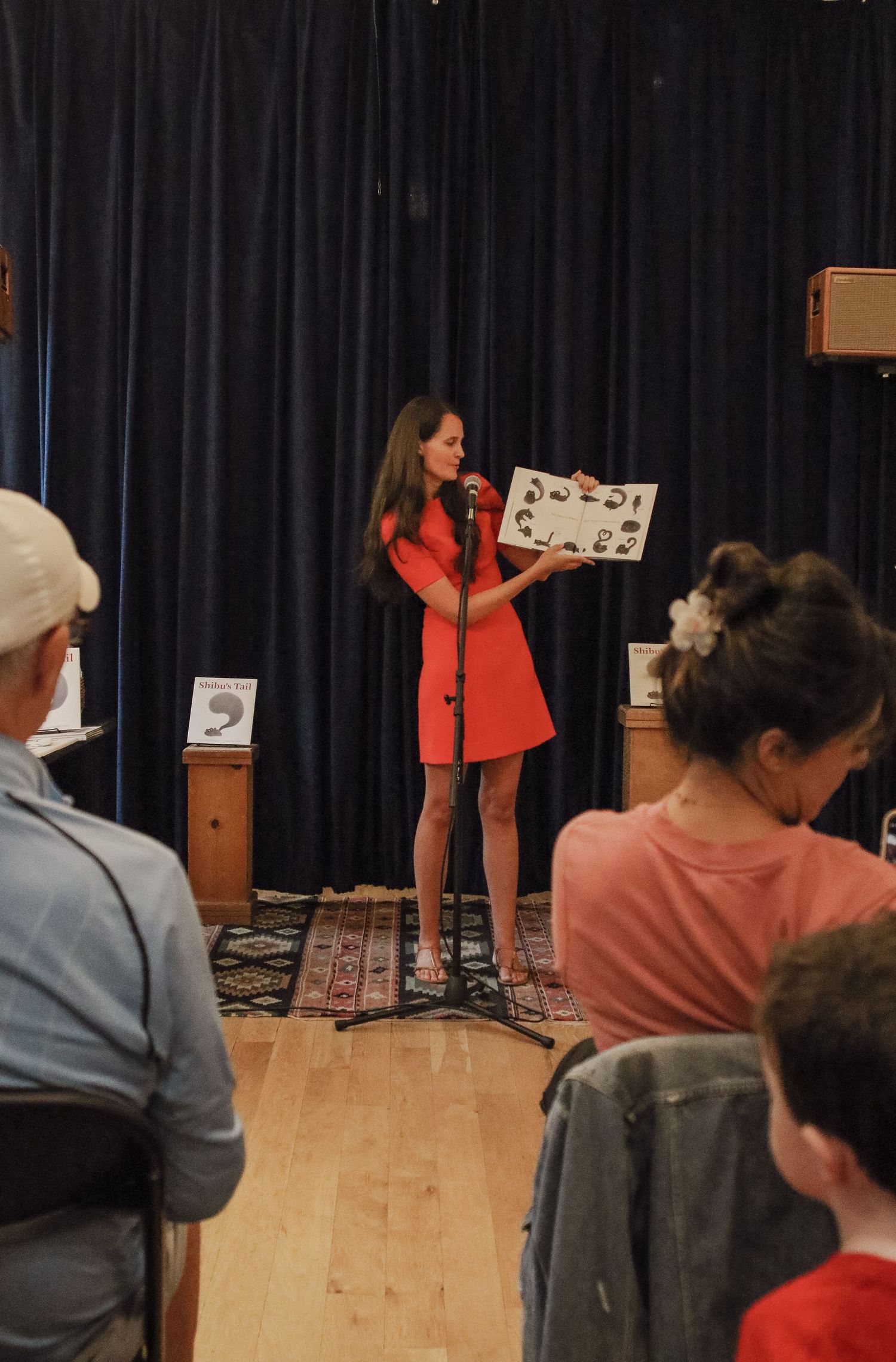 Woman in red dress reading a picture book aloud on stage to a seated audience.