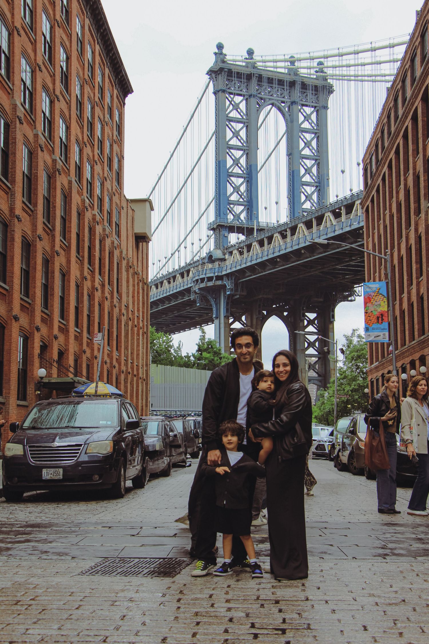 Family posing on cobblestone street with Manhattan Bridge framed between brick buildings in DUMBO Brooklyn.