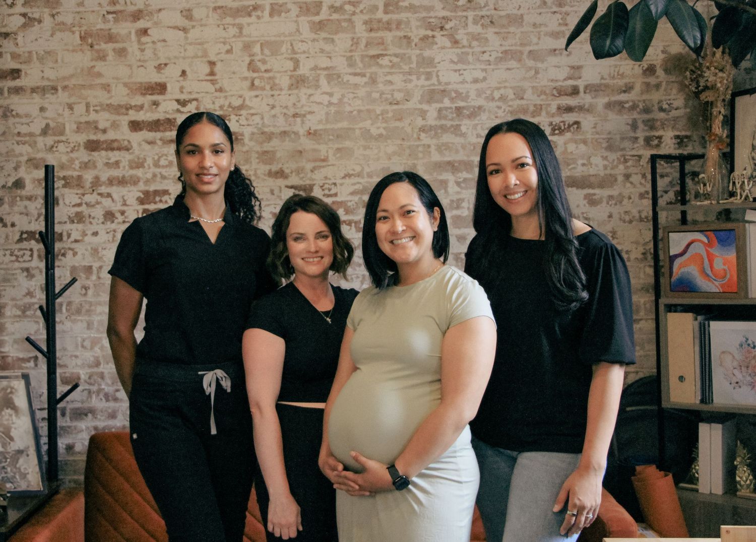Four women posing together indoors, one visibly pregnant cradling her baby bump, against a brick wall.