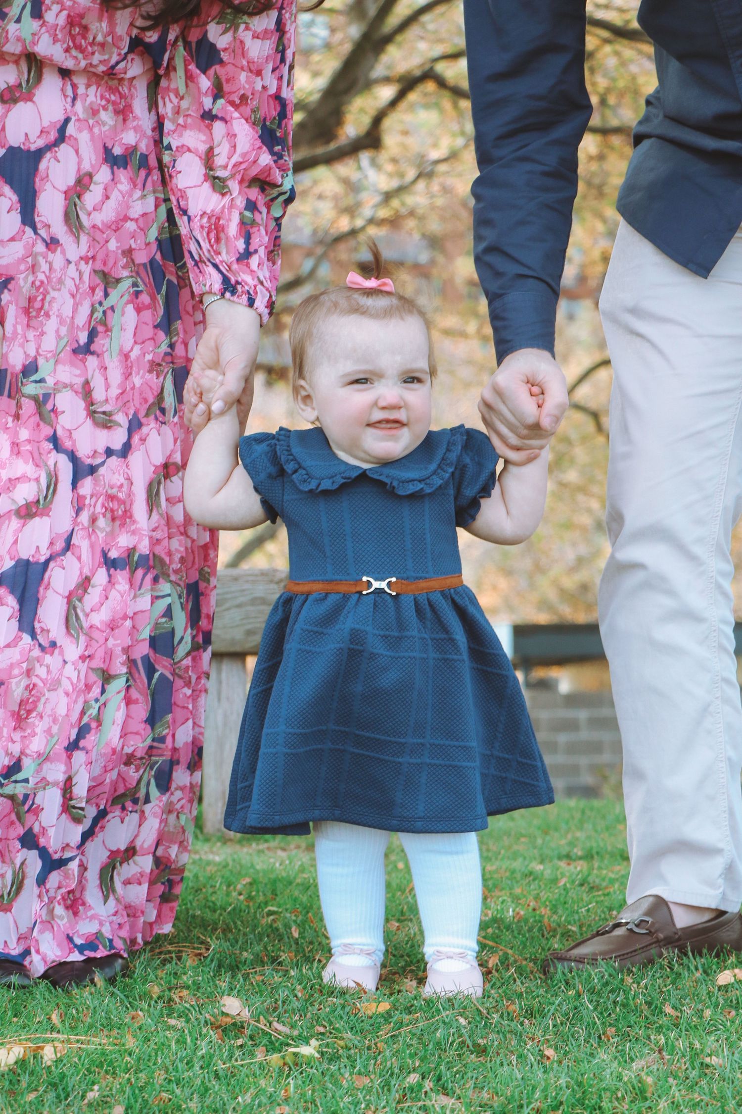 Adorable toddler in navy dress with brown belt stands on grass held by parents during fall photoshoot.