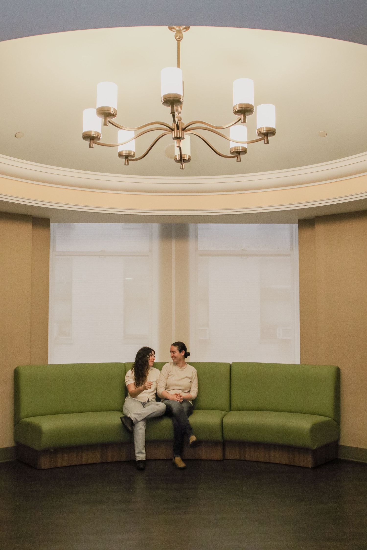 Couple sitting on a curved green sofa beneath an elegant chandelier in a bright room.