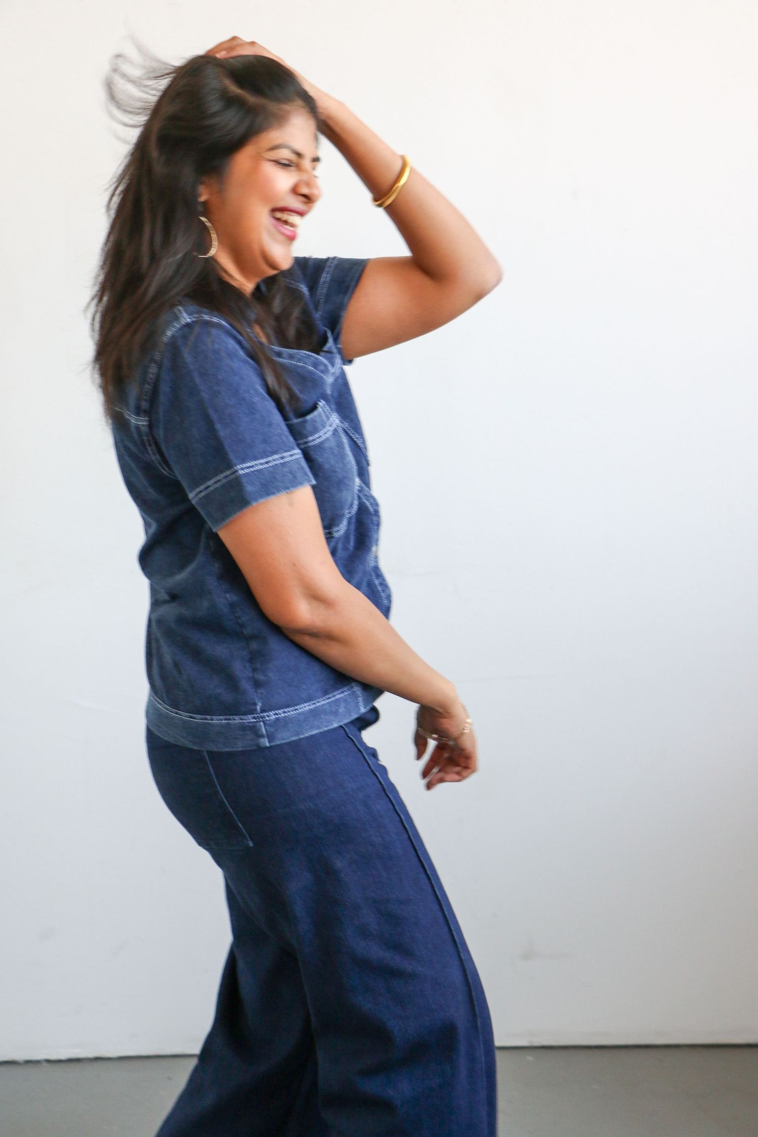 Woman in blue denim outfit laughing and tossing hair playfully against white background.
