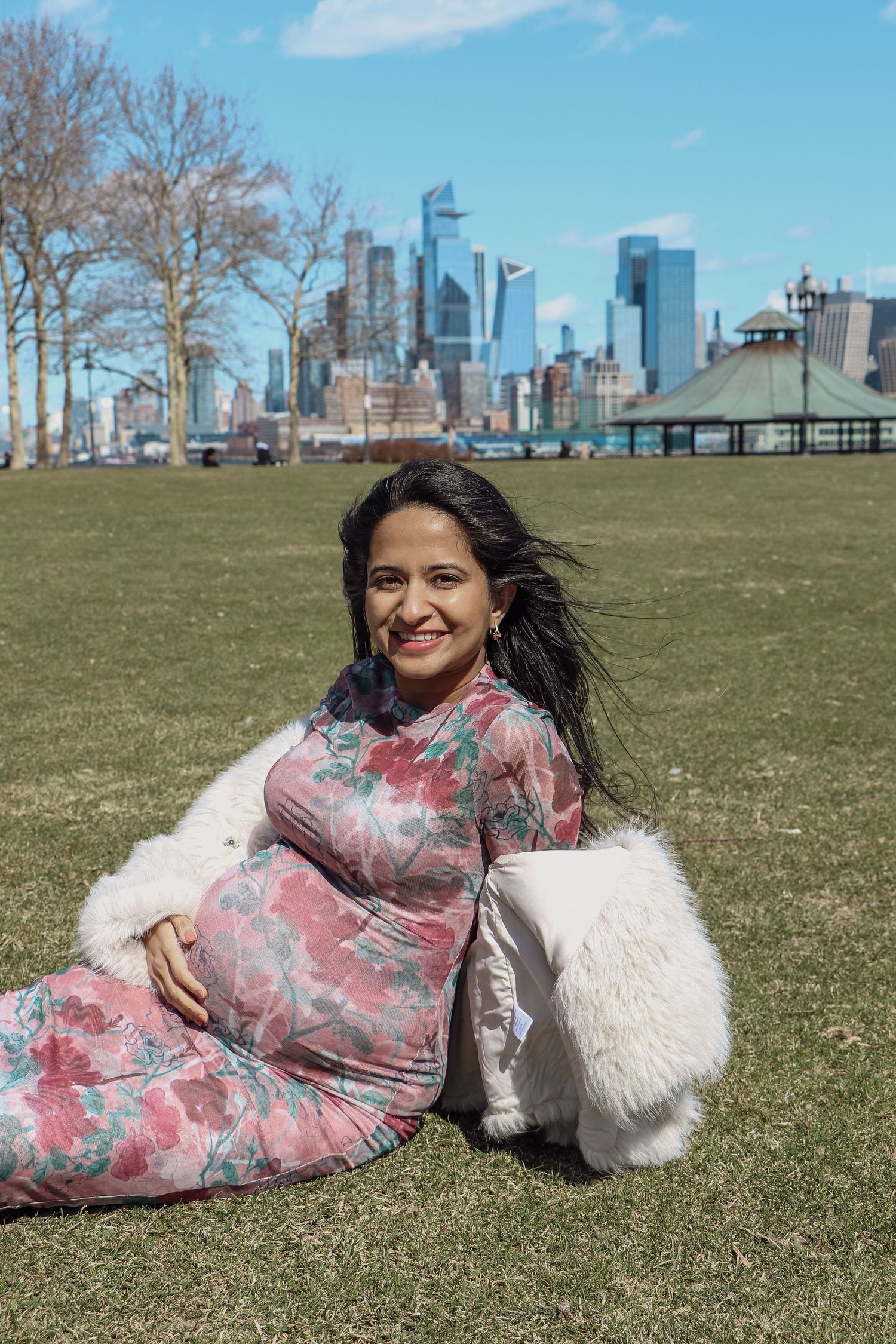 Happy pregnant woman in floral dress sits on green lawn with city skyline in background.